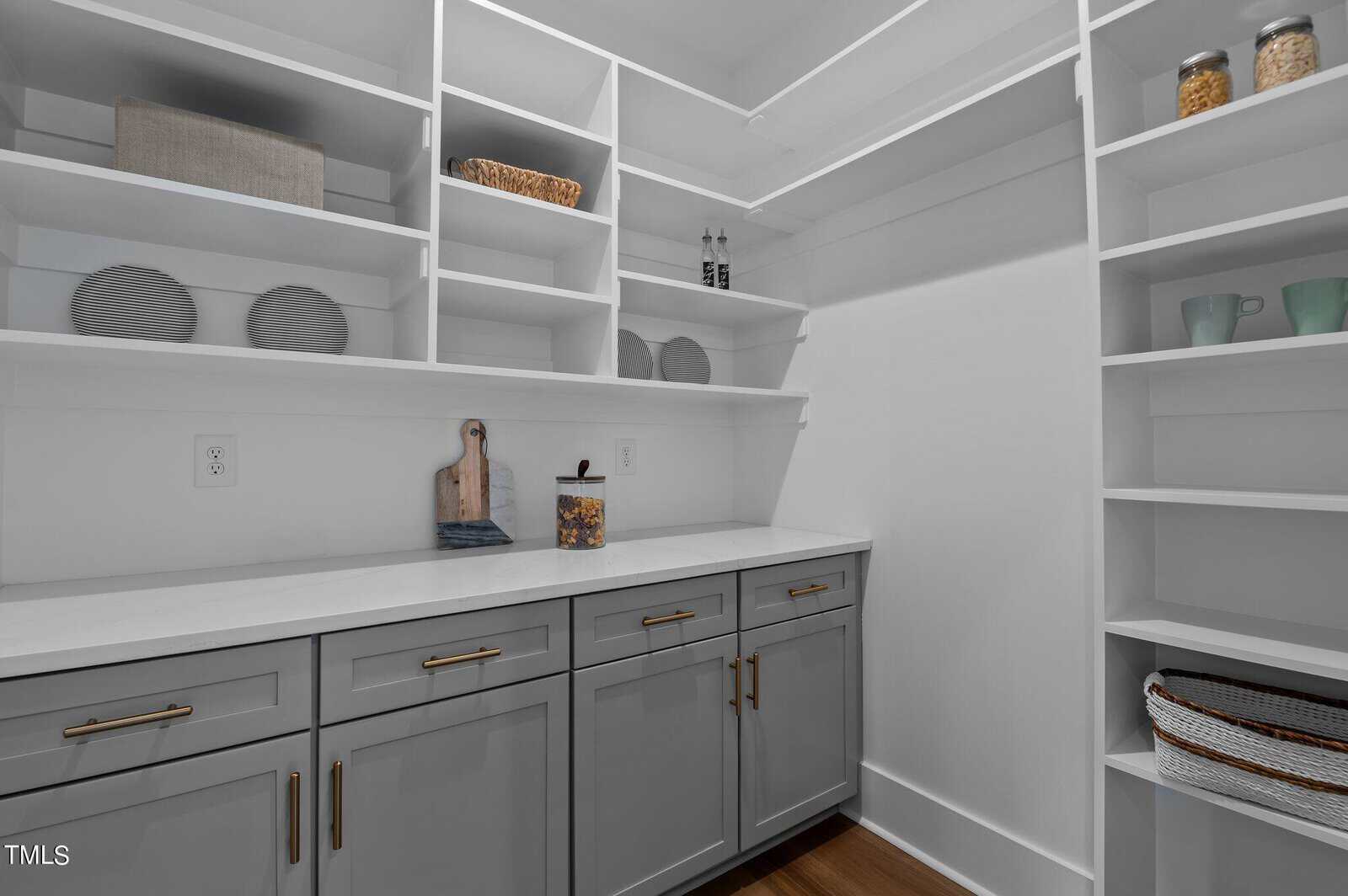 A modern pantry with light gray cabinets, brass handles, white countertops, and open white shelves holding jars, baskets, plates, bowls, cups, and decorative items. The space is neatly organized and well-lit.