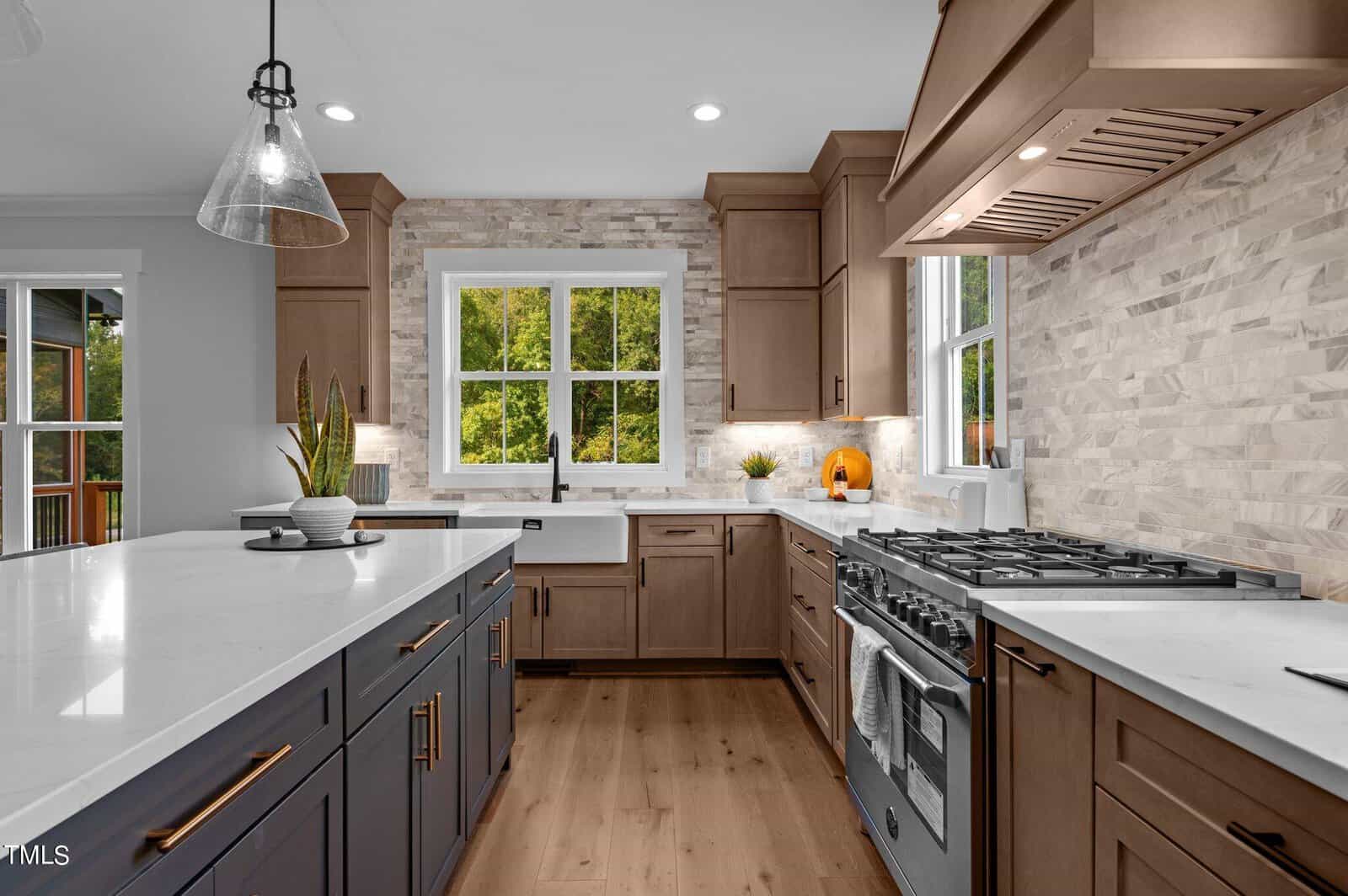 Modern kitchen with wood cabinets, a large island with white countertops, stainless steel appliances, a gas stove, pendant lighting, and large windows overlooking greenery. Light wood floors and pale stone backsplash add warmth.