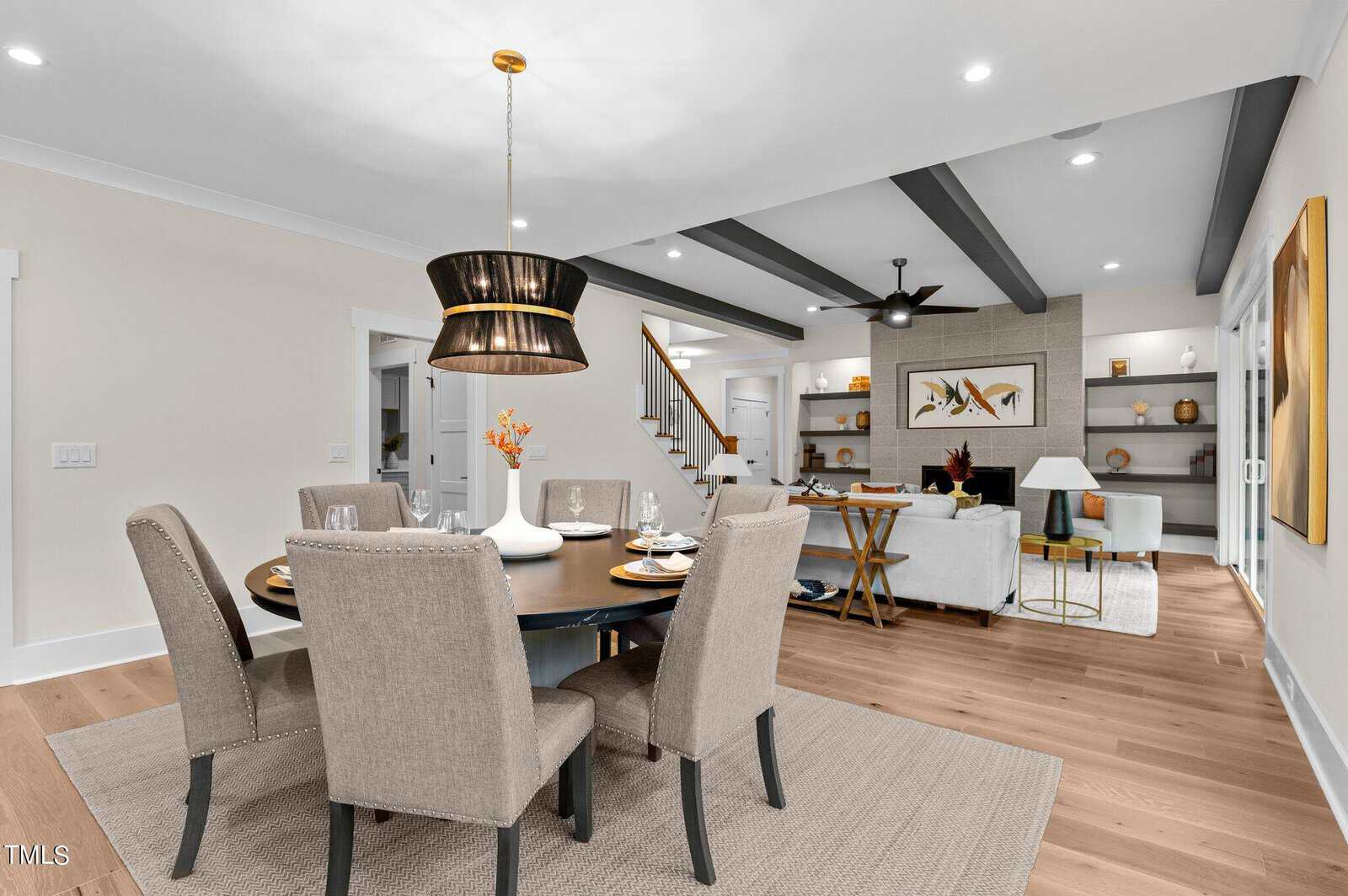 Modern open-concept dining and living area with beige chairs around a dark round table, pendant light above, neutral tones, wood flooring, exposed beams, and built-in shelves in the living room.