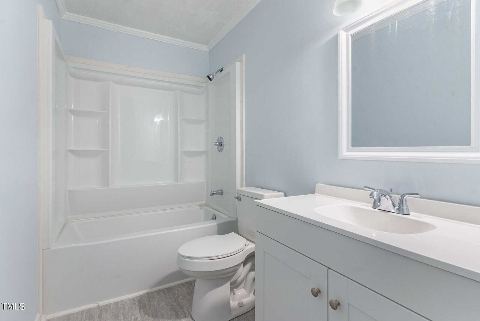 A clean, modern bathroom featuring a white bathtub with an overhead shower, a toilet, and a vanity with a sink and mirror, all set against pale blue walls and gray flooring.