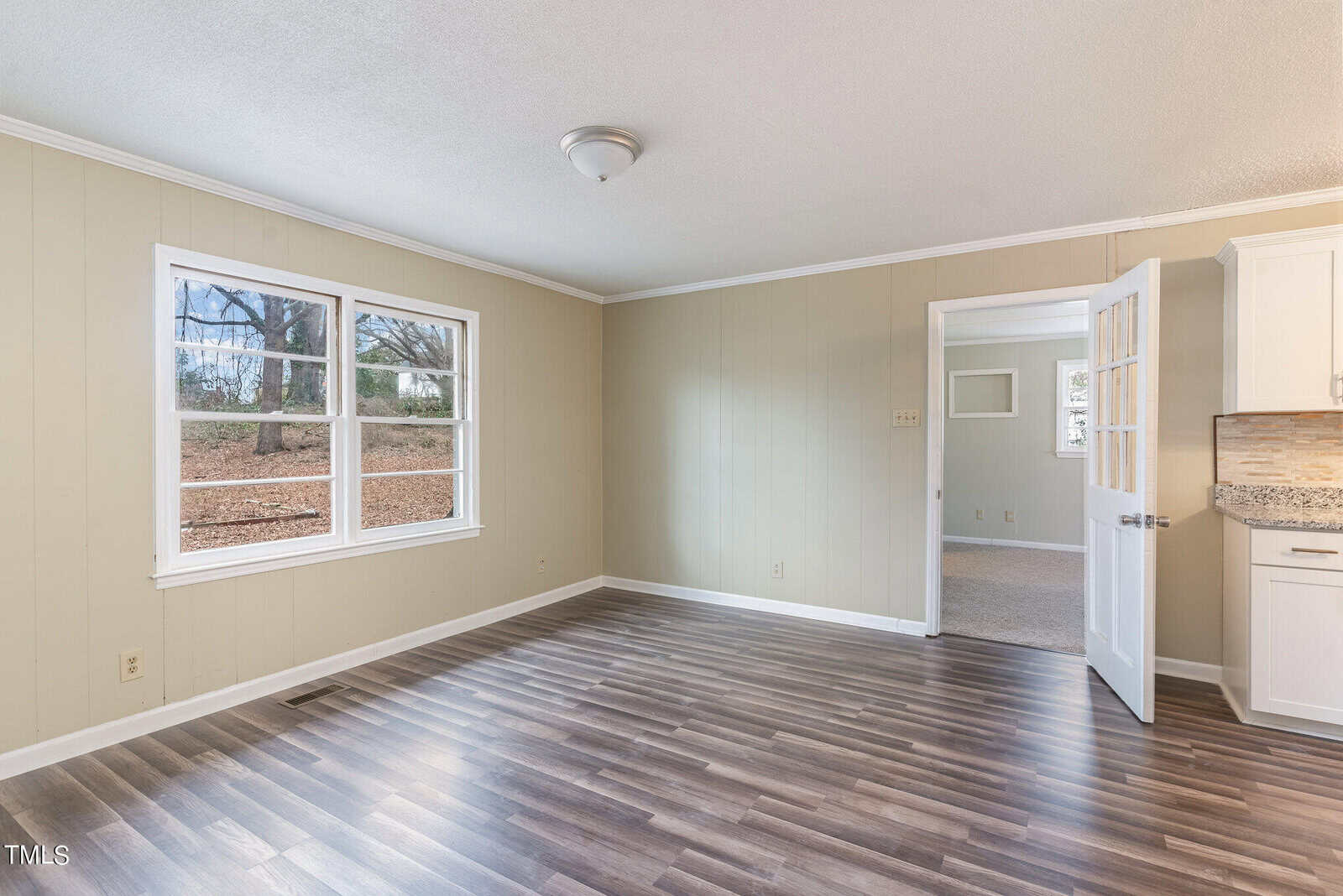 Empty room with beige walls and dark wood flooring, featuring a window with a view of trees and a kitchen entrance visible to the right.