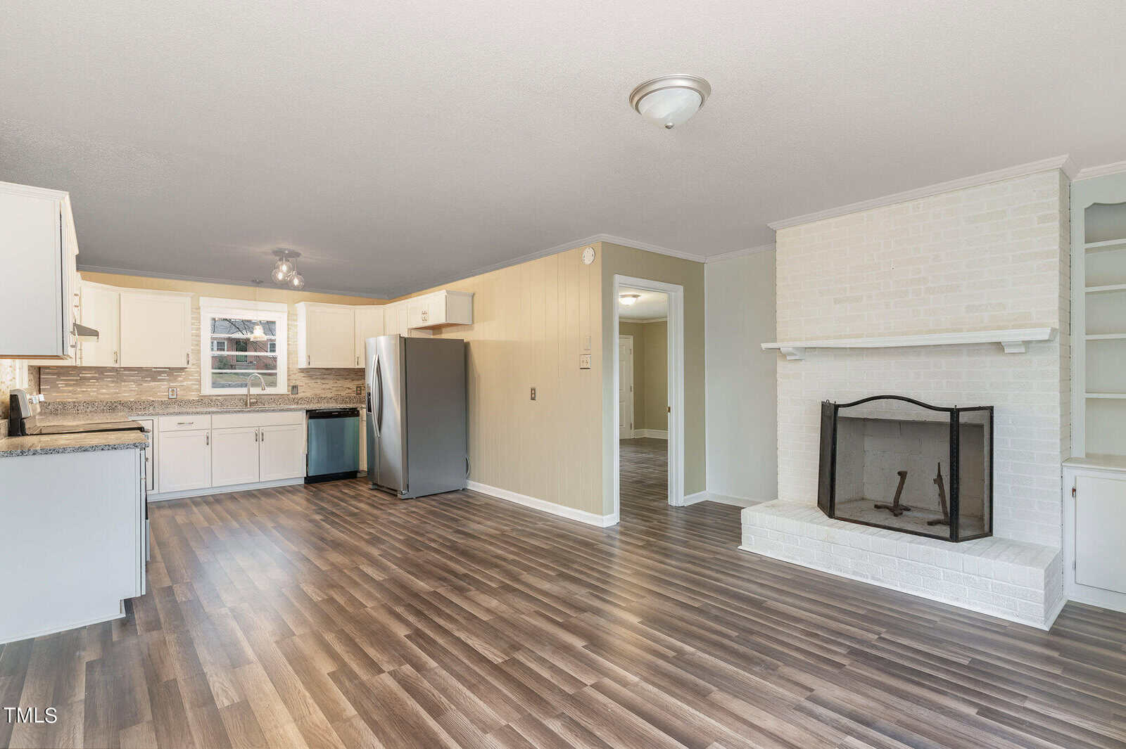 A spacious, well-lit living room featuring glossy wood flooring, a white brick fireplace, built-in shelves, and a view into a kitchen with white cabinets and granite countertops.