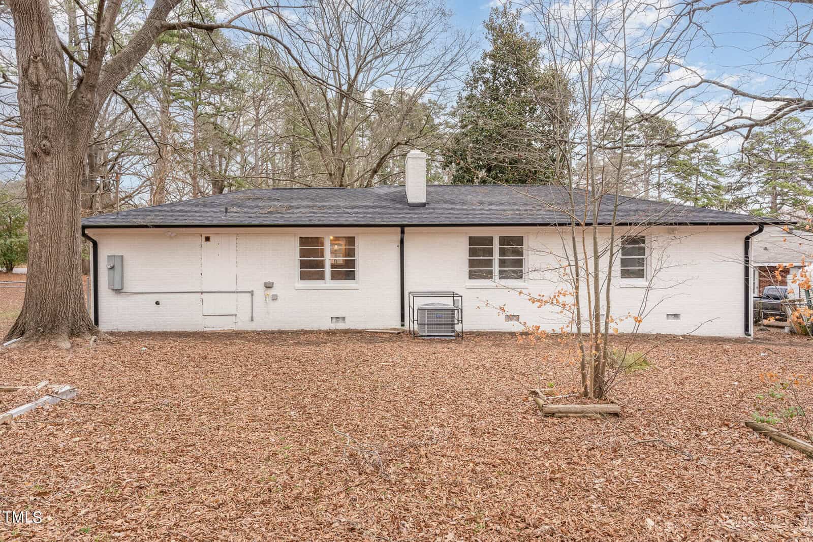 A one-story white brick house with a black shingle roof, several shuttered windows, and a large hvac unit outside, surrounded by a yard covered in fallen leaves.