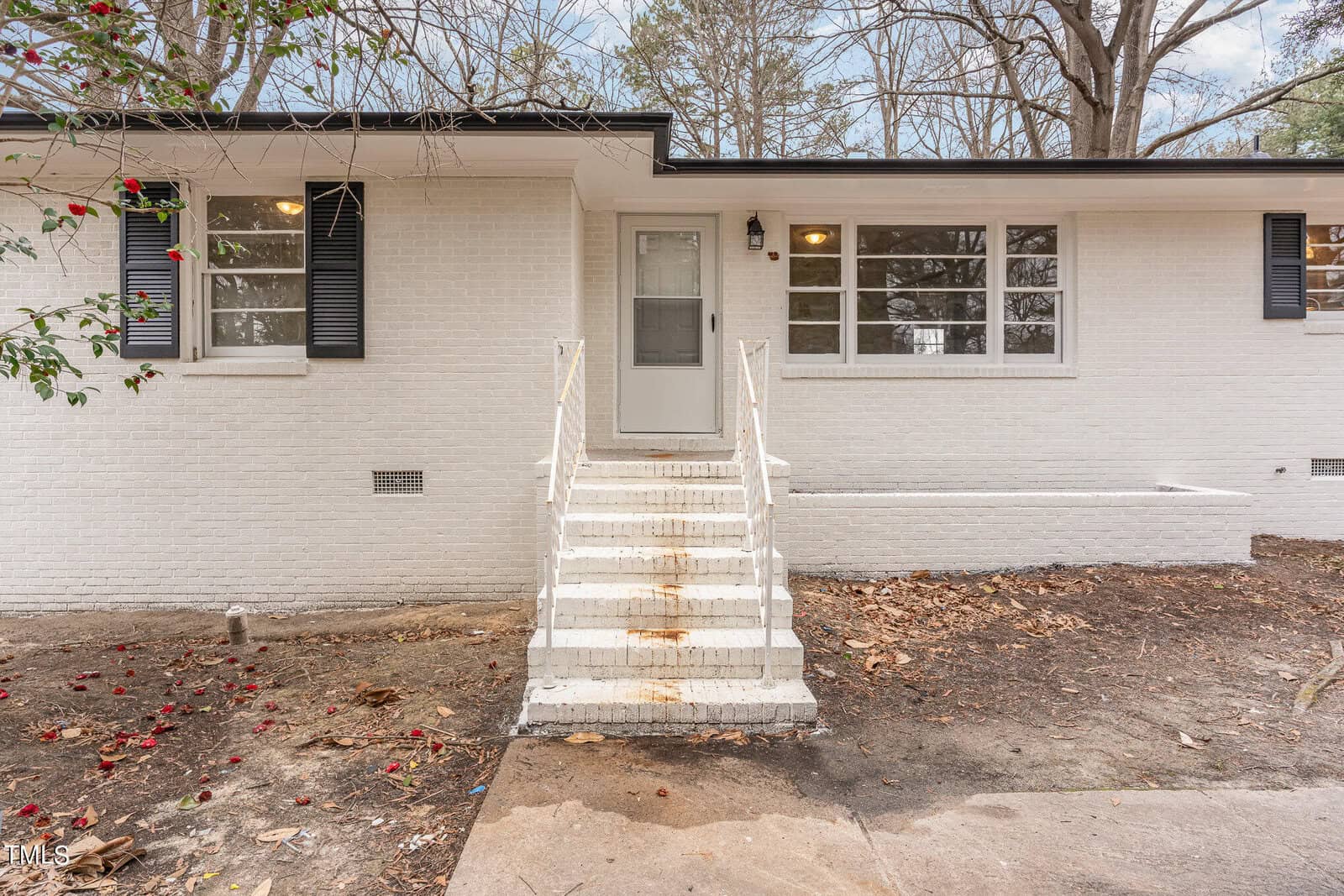 A white brick one-story house with black shutters, featuring a centered front door and a series of white-framed windows. a small staircase with white railings leads up to the entrance.