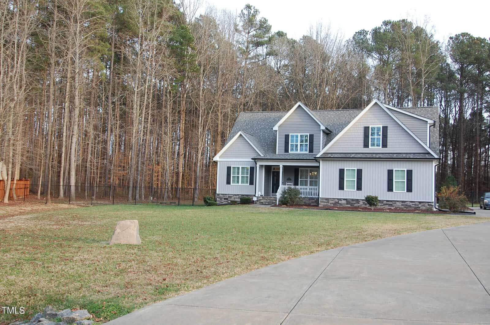 A two-story gray house with a sloped roof, surrounded by a wooded area. The home features a small porch, manicured lawn, and a long driveway. Trees with no leaves are visible in the background, indicating a winter or early spring setting.