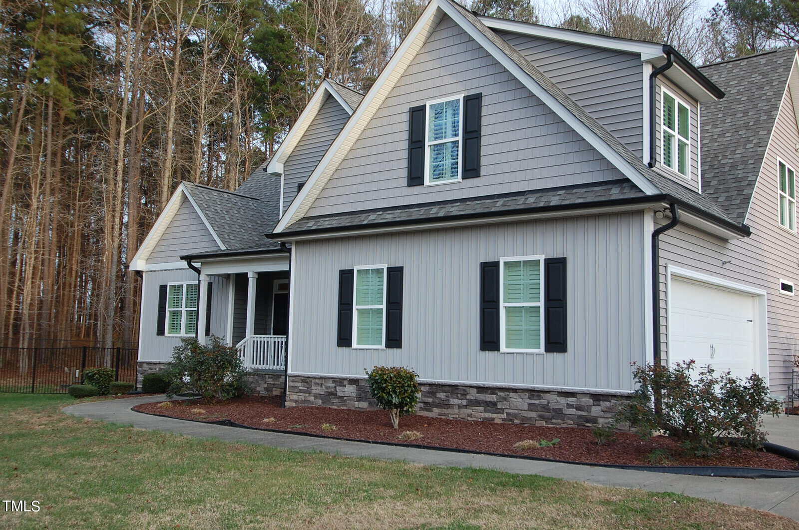 A gray two-story house with a gable roof and green shutters. The front yard features neatly manicured shrubs and a curved driveway. A wooded area is visible in the background.