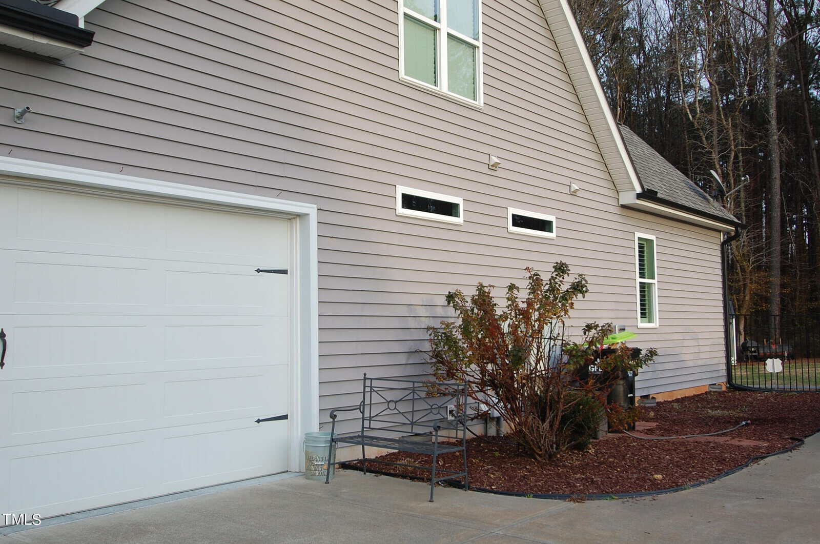 Side view of a house with pale gray siding and a white garage door. A small garden area has bushes and a metal bench on a paved area. Trees are visible in the background.