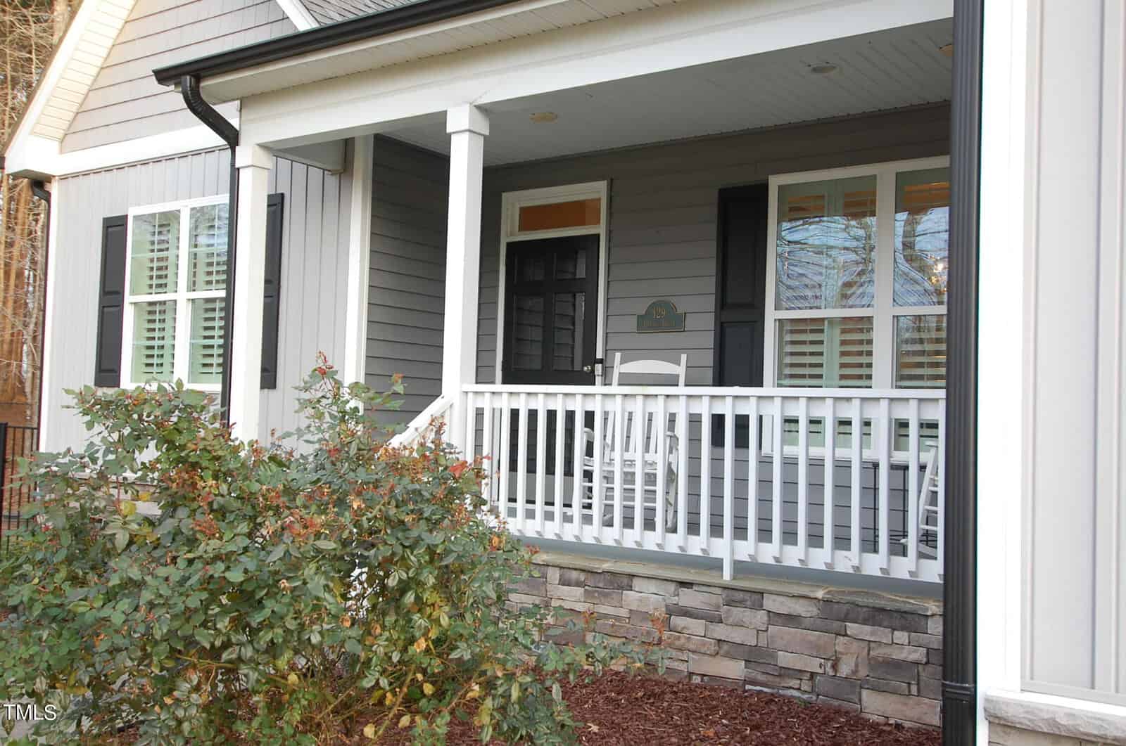 A cozy front porch with a white railing and two rocking chairs. The siding is gray with white trim, and there's a stone accent on the lower half. A small bush with green leaves is in the foreground.