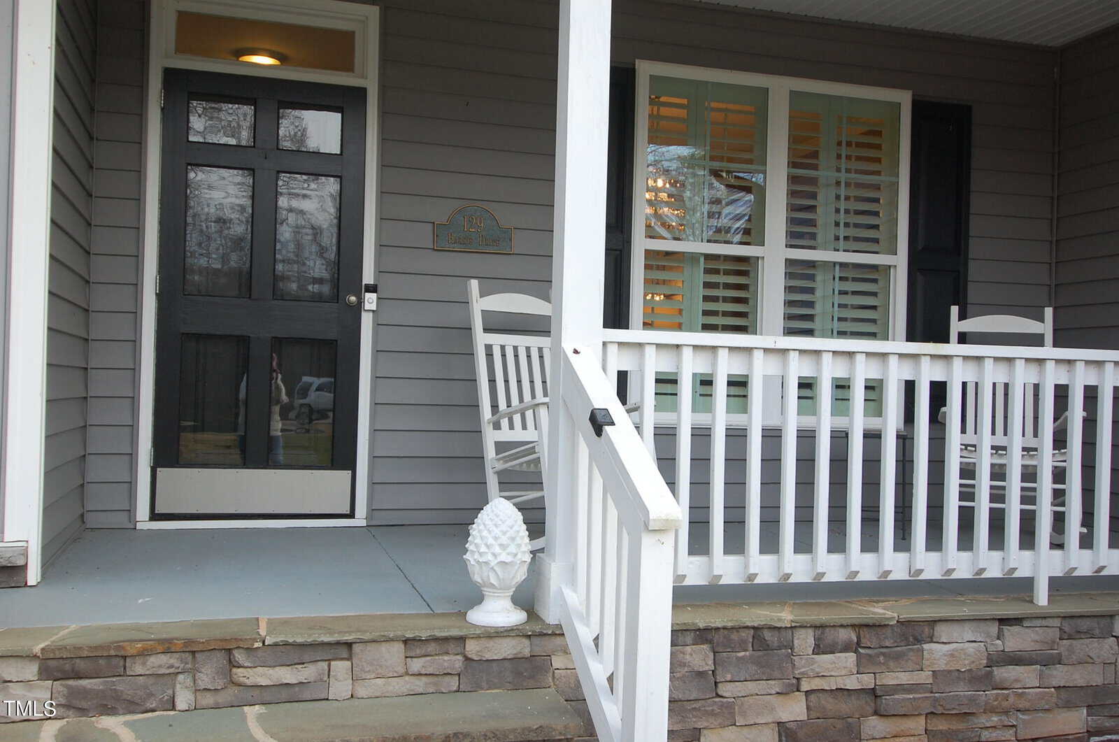 A cozy house entrance with a closed dark green door and front porch. Two white rocking chairs sit on the porch beside a white railing. A decorative pineapple ornament is on a stone step. Shuttered windows are on the right.