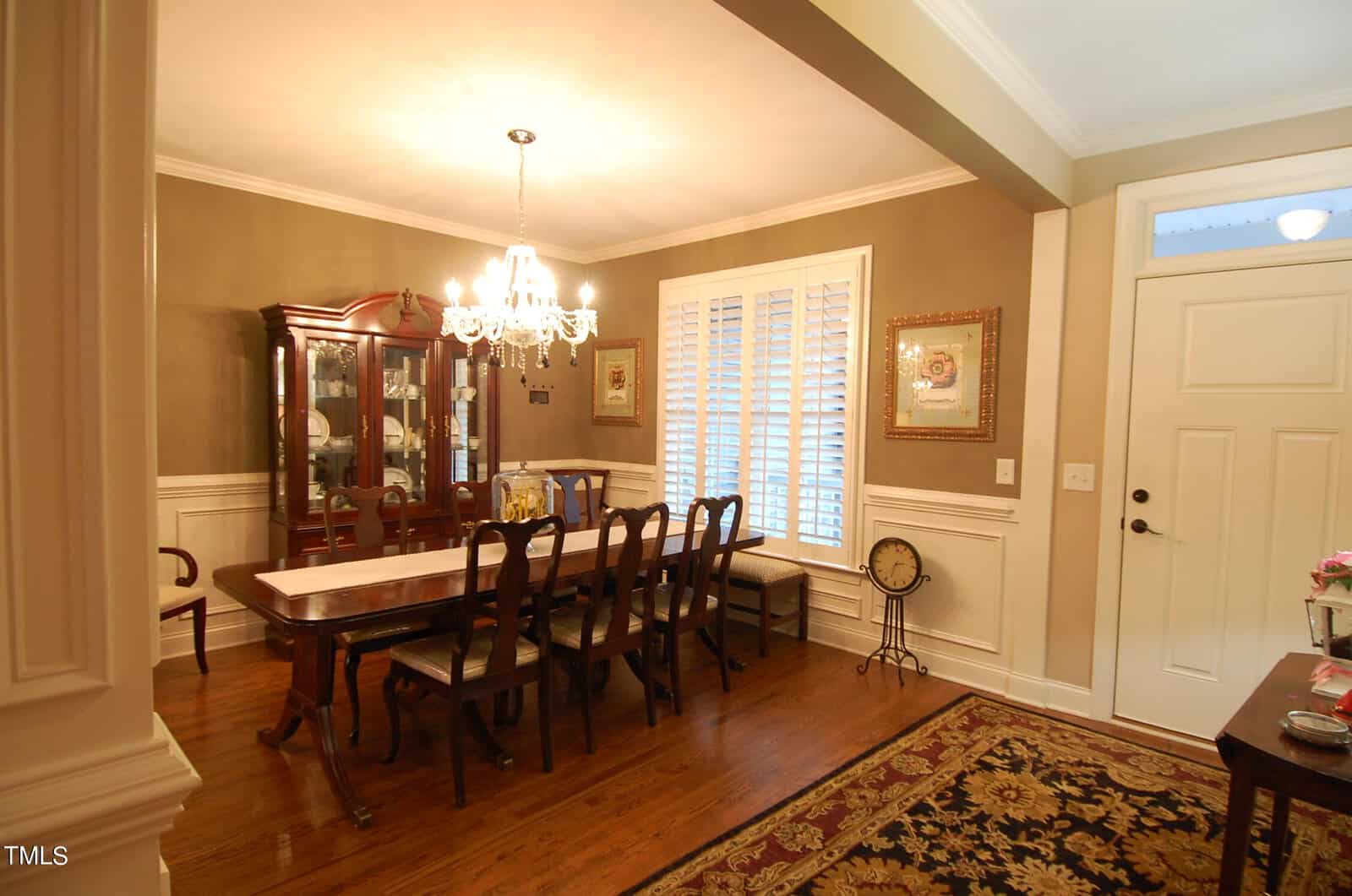 Elegant dining room with a long wooden table and eight chairs, a chandelier overhead, and a china cabinet in the corner. The room has hardwood floors, beige walls with wainscoting, and a large window with blinds. A floral rug is near the entrance.