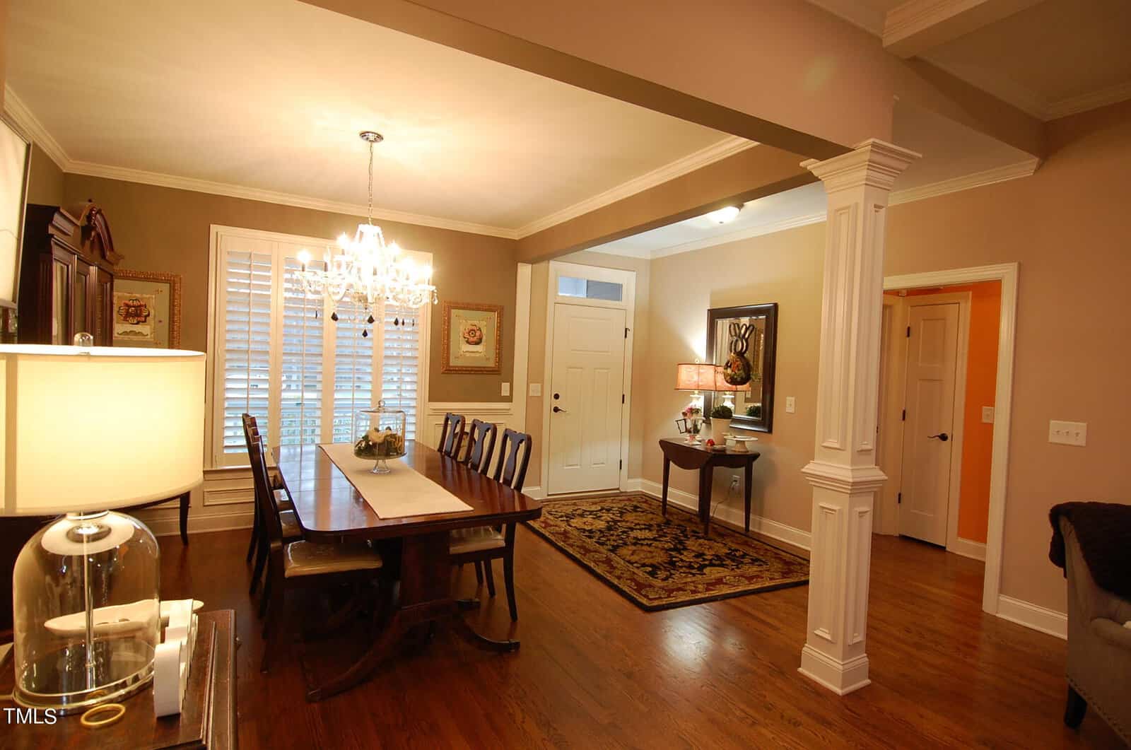A warm and welcoming dining room with a large wooden table surrounded by chairs. A chandelier hangs overhead, and a side table with a lamp sits near an entrance door. Hardwood floors and soft lighting create a cozy atmosphere.