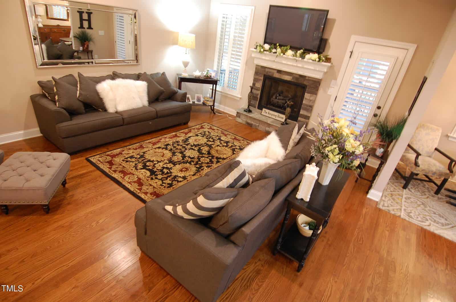 A cozy living room features two gray sofas with pillows, a patterned rug, and a fireplace adorned with floral decorations. A wall-mounted TV is above the fireplace. Hardwood floors and a small ottoman complete the inviting space.
