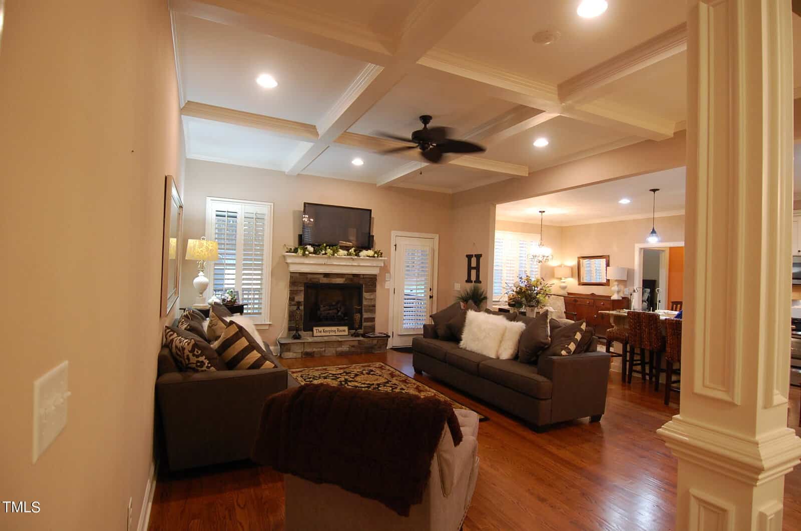 A cozy living room with a warm, earthy color scheme. It features two sofas, a stone fireplace with a mounted TV above, and recessed lighting. The dining and kitchen areas are visible in the background. Wood floors add to the inviting atmosphere.