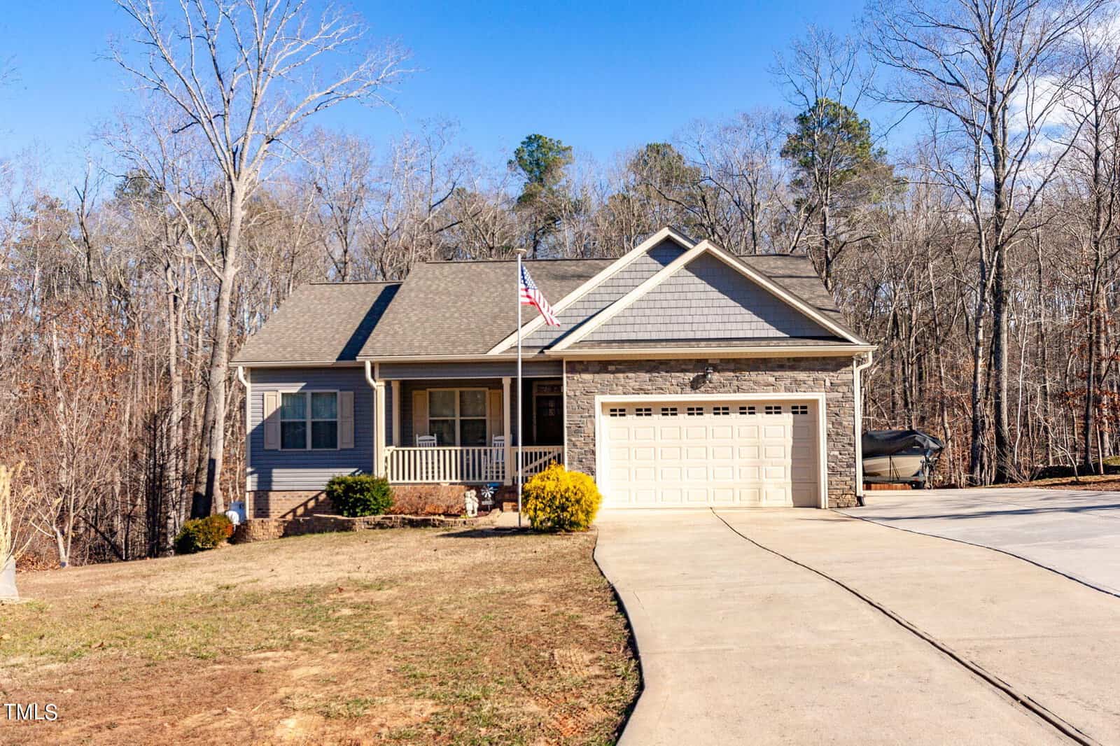 A single-story gray house at 2496 Golden Forest Drive in Franklinton features a front porch, two-car garage, and an American flag. The home is surrounded by leafless trees and a large driveway under a clear blue sky.