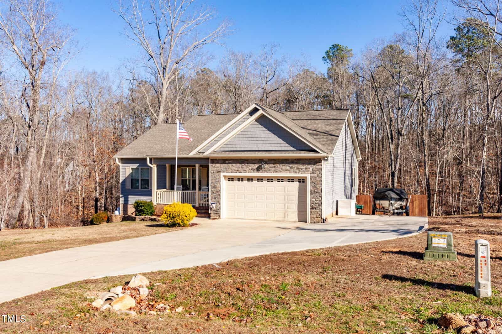 Single-story house with a front porch, two-car garage, American flag, and a barbecue grill on the driveway. The yard has sparse grass and bushes, and the home is set against a backdrop of tall, leafless trees.