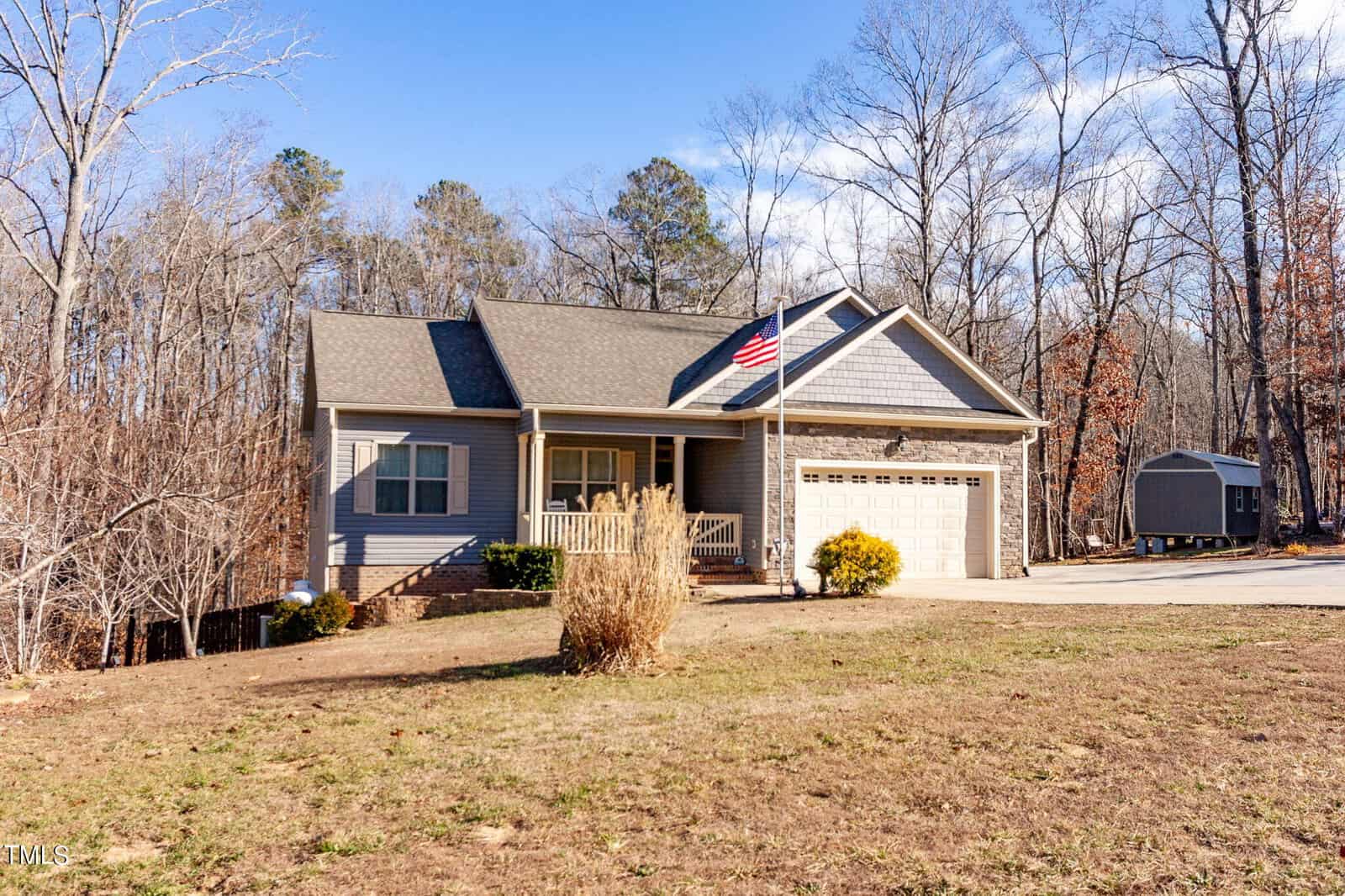 A single-story gray house with a front porch, an American flag, and a double garage is surrounded by trees and a large yard under a clear blue sky. A small shed is visible in the background.