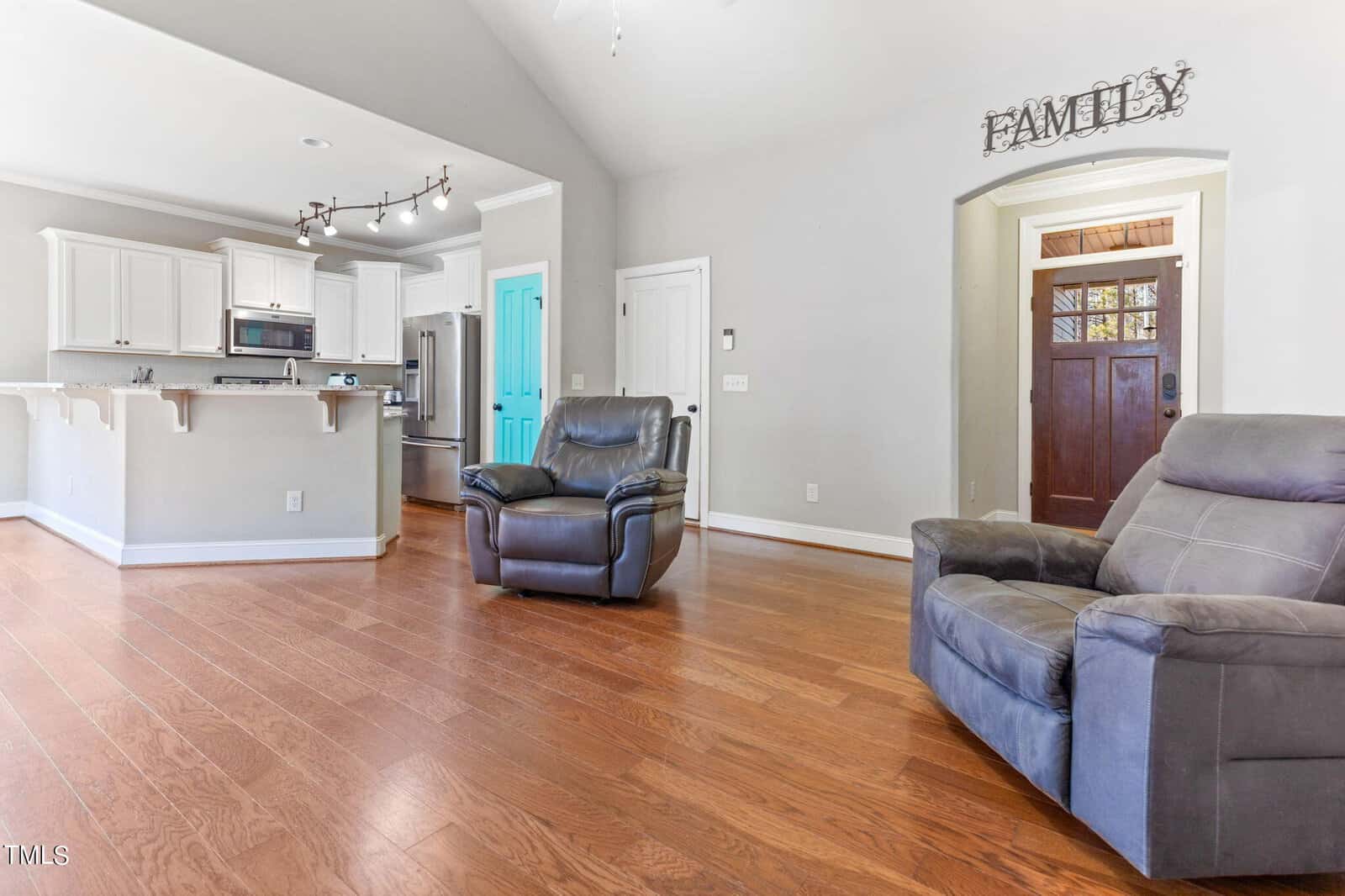 A bright living room with wood floors, two gray recliners, a kitchen with white cabinets, a blue pantry door, and a wooden front door beneath a sign that reads "FAMILY.