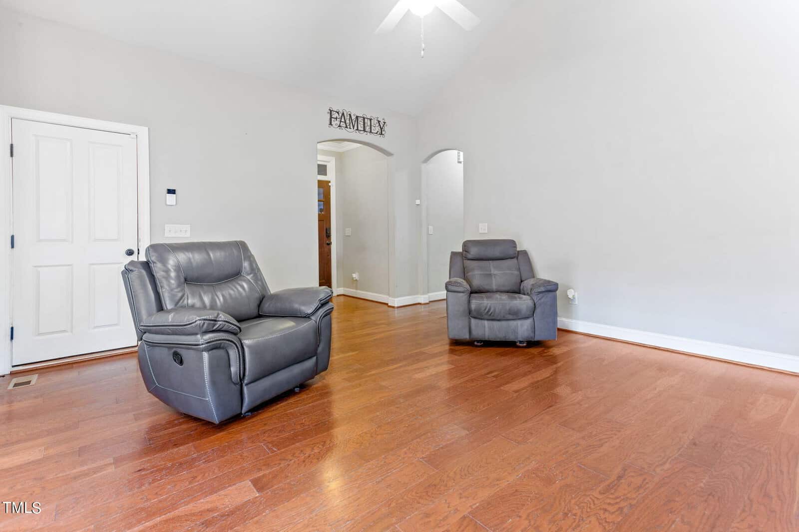 A living room with two gray leather recliners on hardwood floors, light gray walls, a white door, and an arched doorway with a "FAMILY" sign above it. The room is simple and uncluttered.