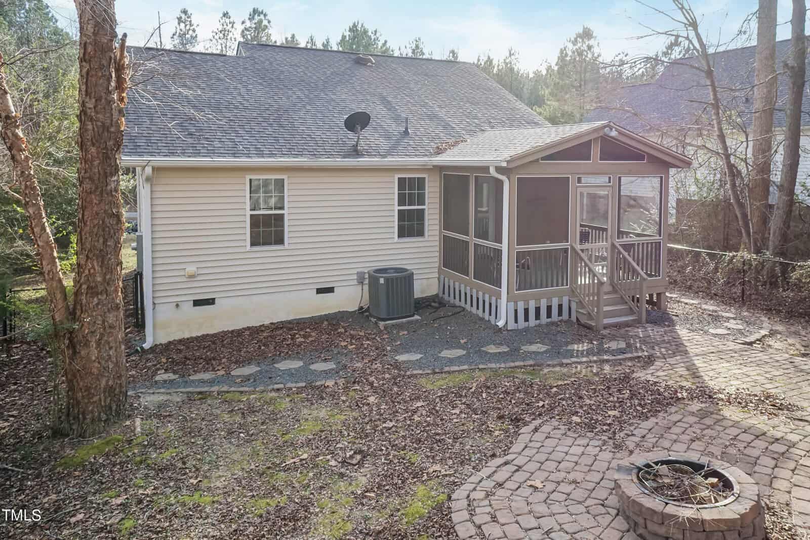 A backyard view of a single-story house with a screened porch. The yard features stepping stones leading to a brick patio with a fire pit. Surrounded by trees, it is a serene outdoor space.