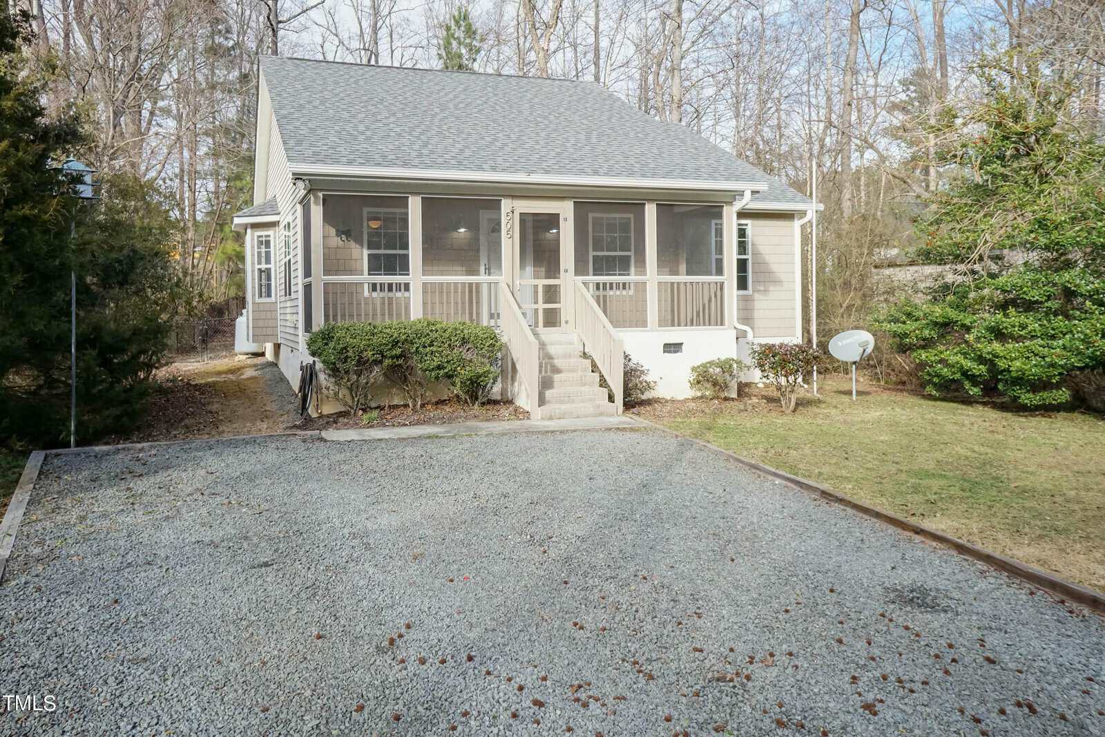 A small, single-story house with a gray roof and screened front porch. A staircase leads to the porch. The yard is bordered by trees, and there's a gravel driveway in the foreground and a satellite dish on the right.