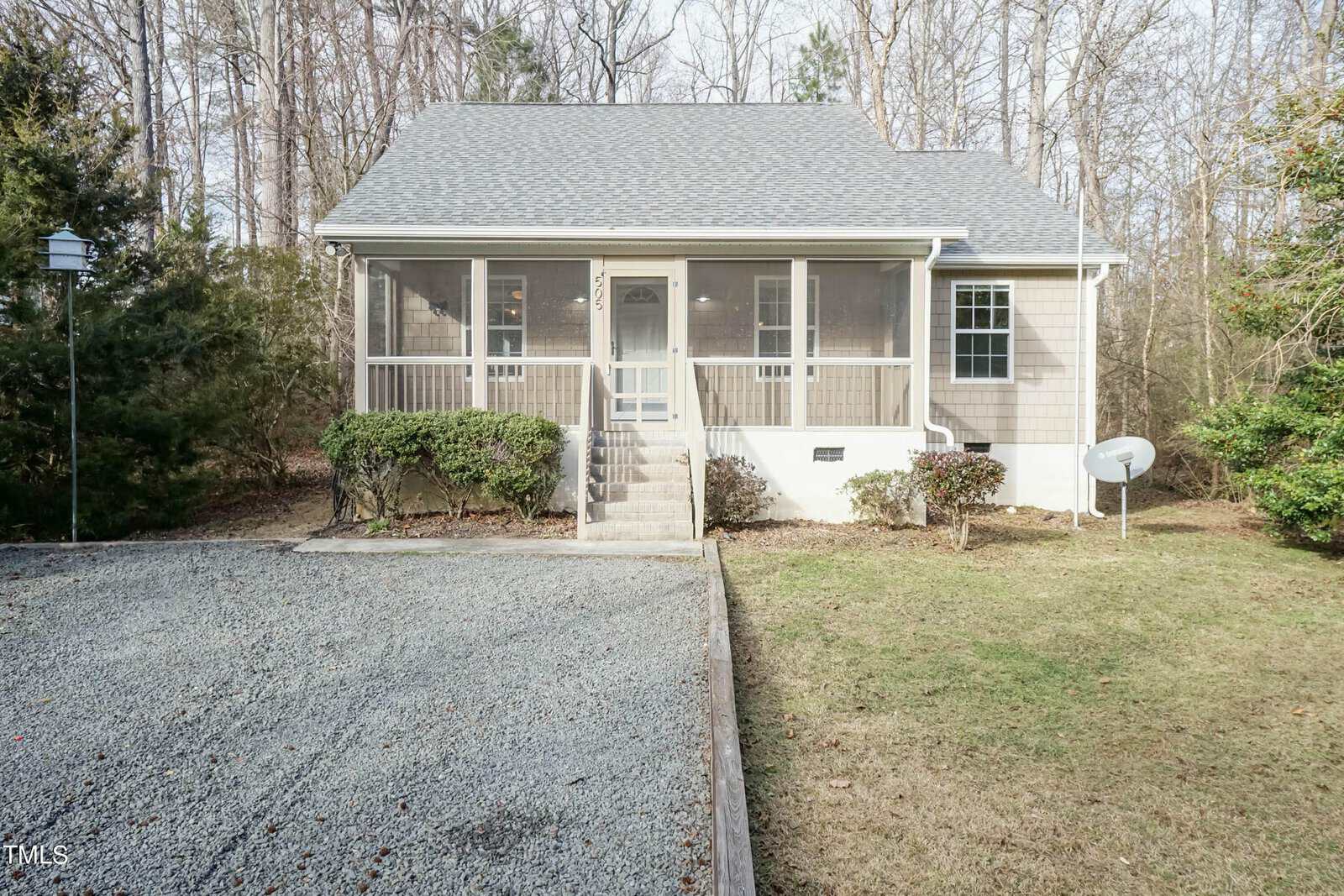 A small house with light gray siding and a screened front porch is surrounded by trees. The house has a gabled roof, a gravel driveway, and a satellite dish on the right. Steps lead up to the porch, and shrubs line the path.