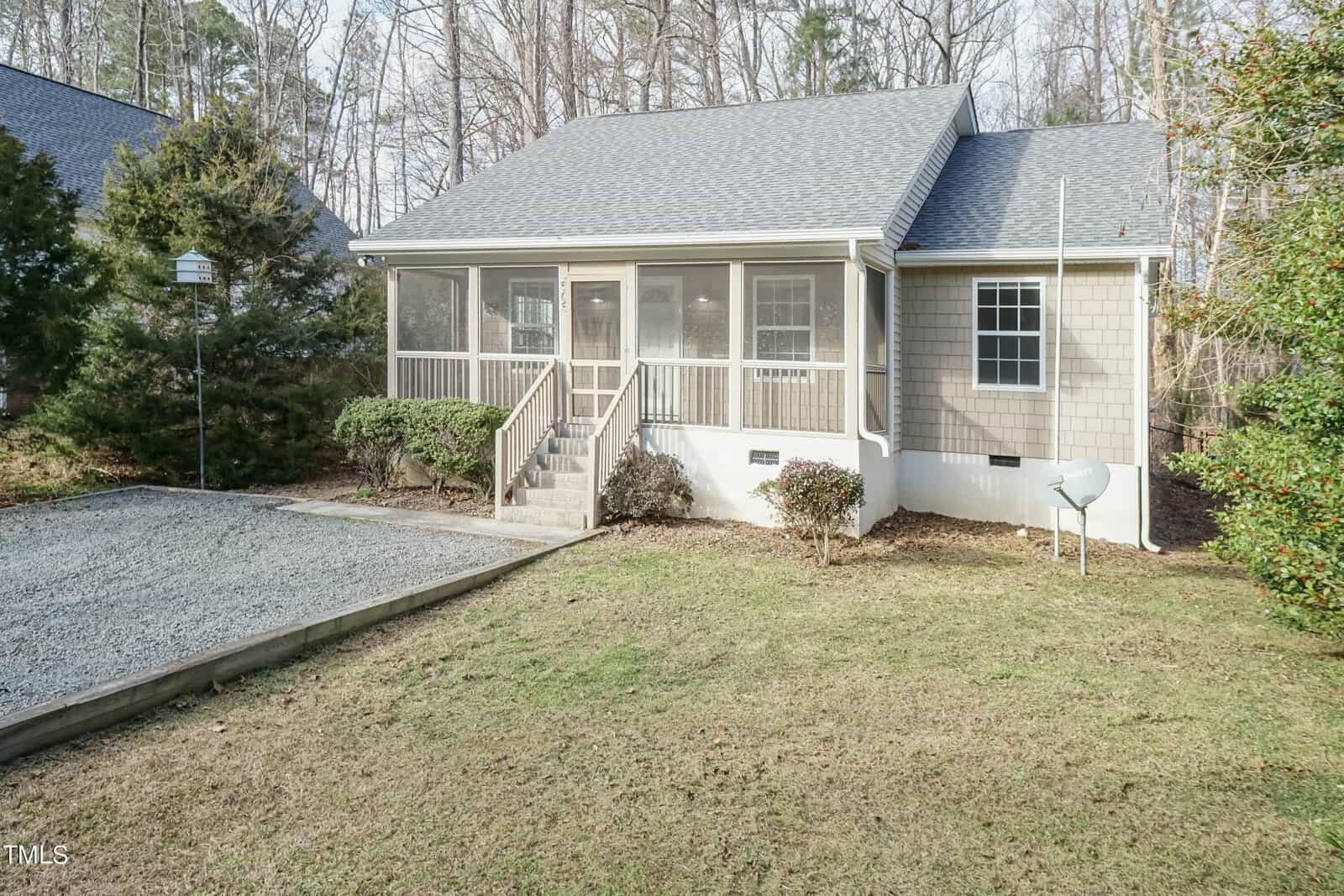 A cozy, single-story house with a screened-in front porch, nestled among trees. The exterior is a mix of siding and brick, with a gravel driveway and a well-maintained lawn in front.