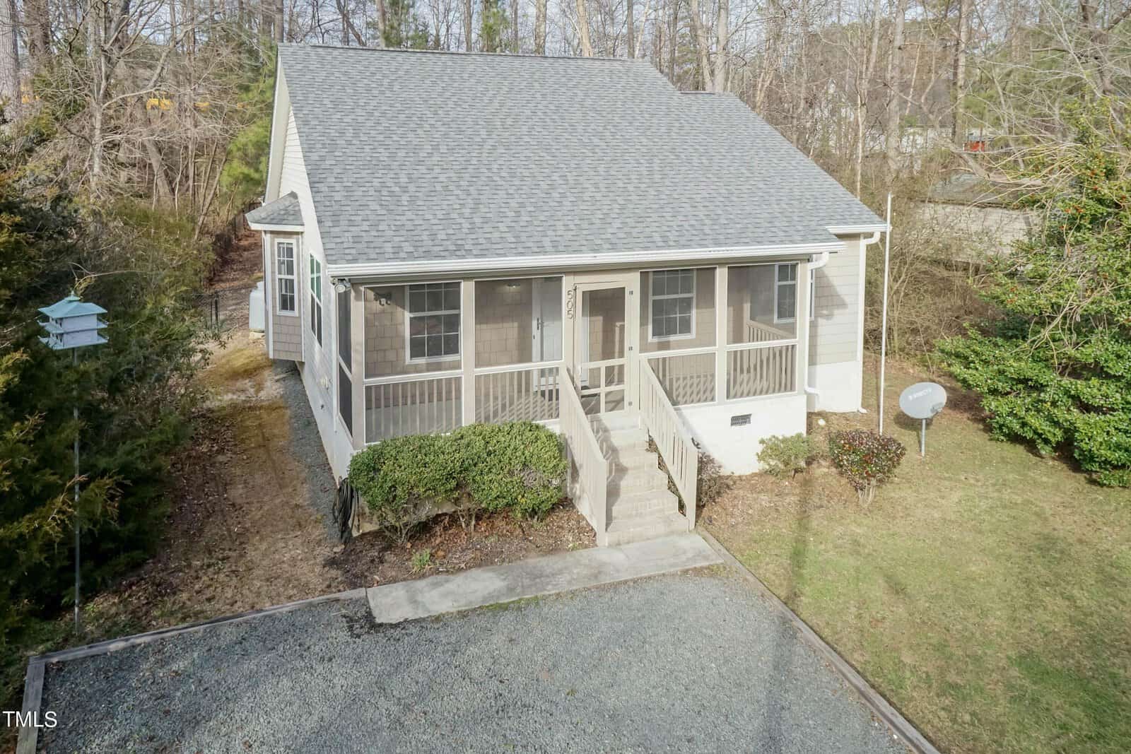 A small, single-story house with a gray roof and white siding, featuring a screened porch and a front staircase. The yard is grassy with a few bushes. A gravel driveway is in front, and there are trees in the background.