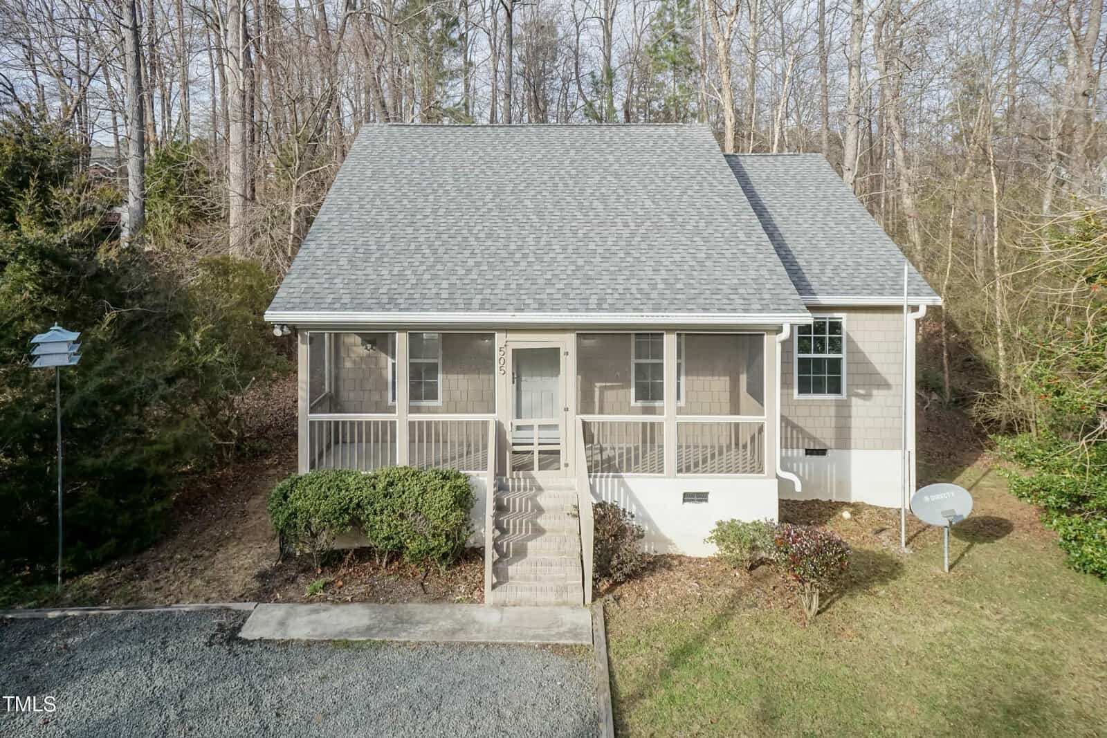 A quaint two-story house at 505 Crescent Heights Drive, featuring a gray roof and beige siding, sits in a wooded area of Creedmoor. It boasts a screened front porch with steps leading to the door. A gravel driveway lies in the foreground, with a satellite dish on the right.