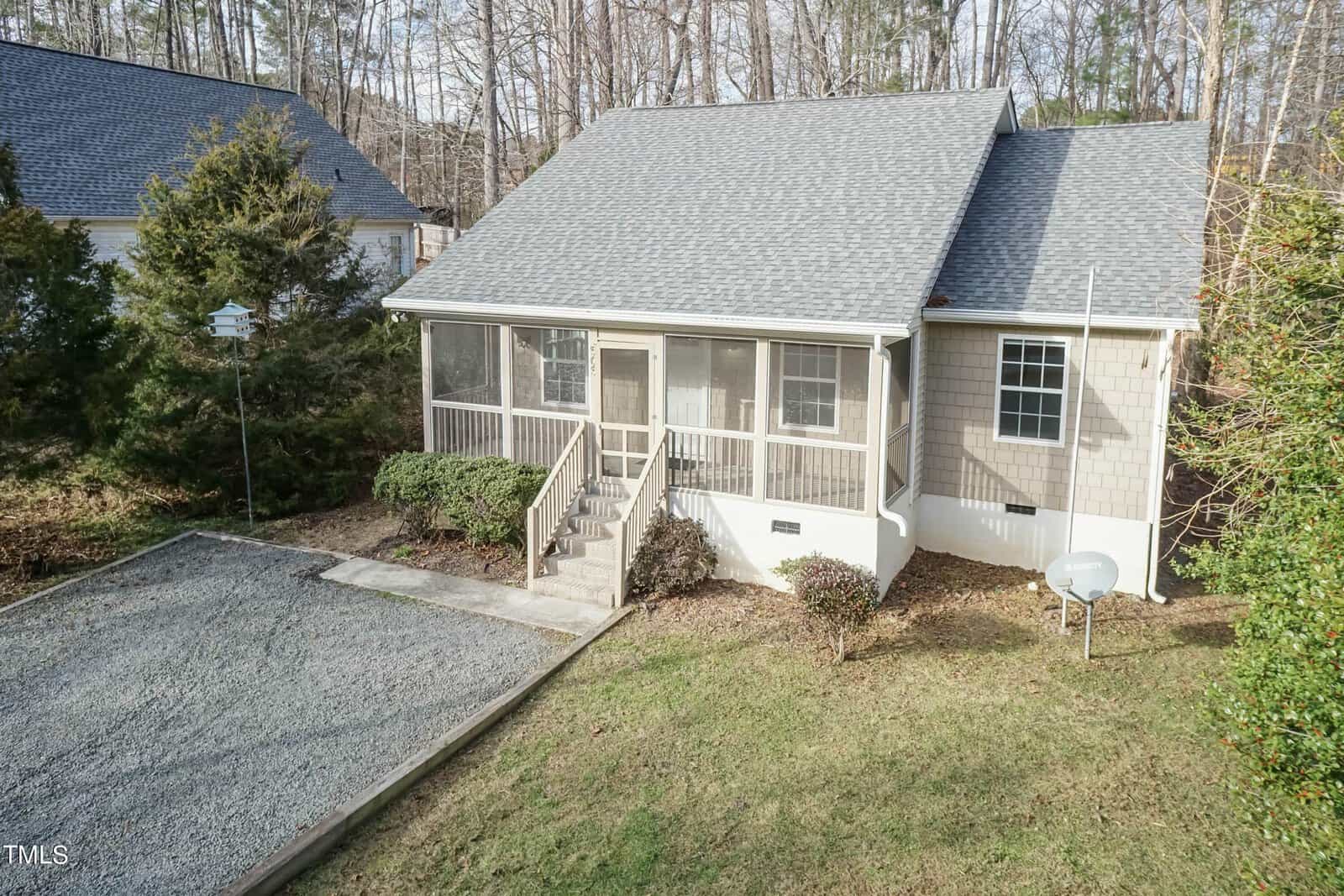 A small beige house with a gabled roof and a screened-in front porch, surrounded by trees. A gravel driveway leads up to the front steps, and there's a satellite dish on the side of the house. The area is wooded and serene.