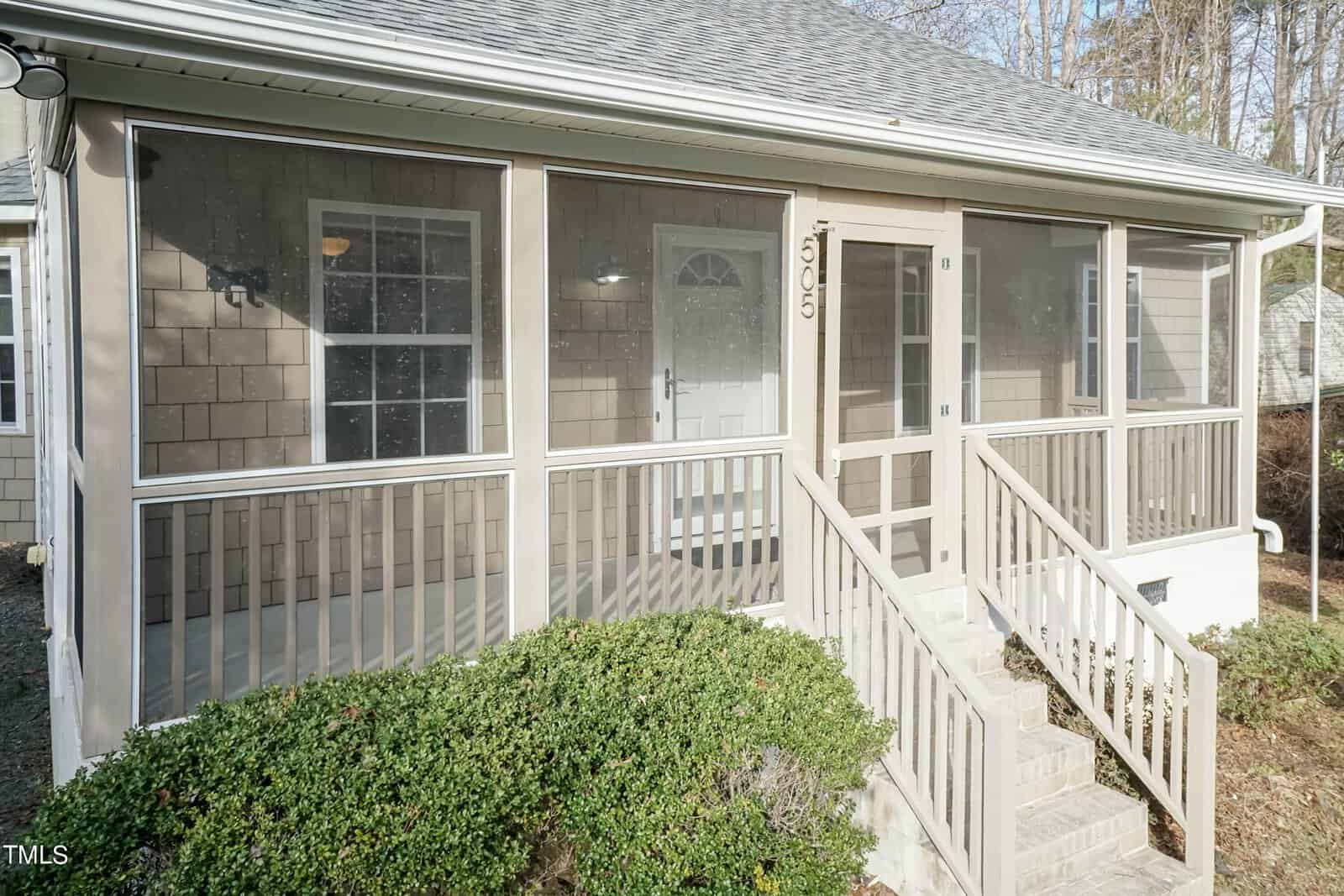 A small house with shingle siding featuring a screened-in front porch. The porch has a door and simple railing, with steps leading up to it. Bushes border the front of the porch. The house number "1015" is displayed above the door.