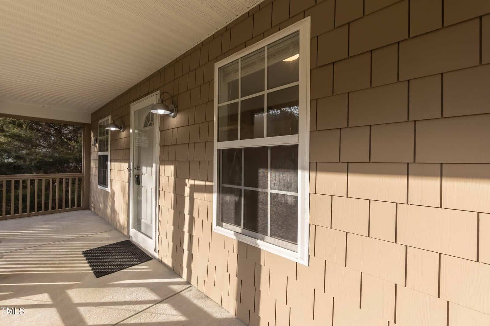 A covered porch with beige shingle siding, featuring two windows and a door with a small black mat. Two wall-mounted lights are positioned above the door and window, and the porch is surrounded by a wooden railing with trees in the background.