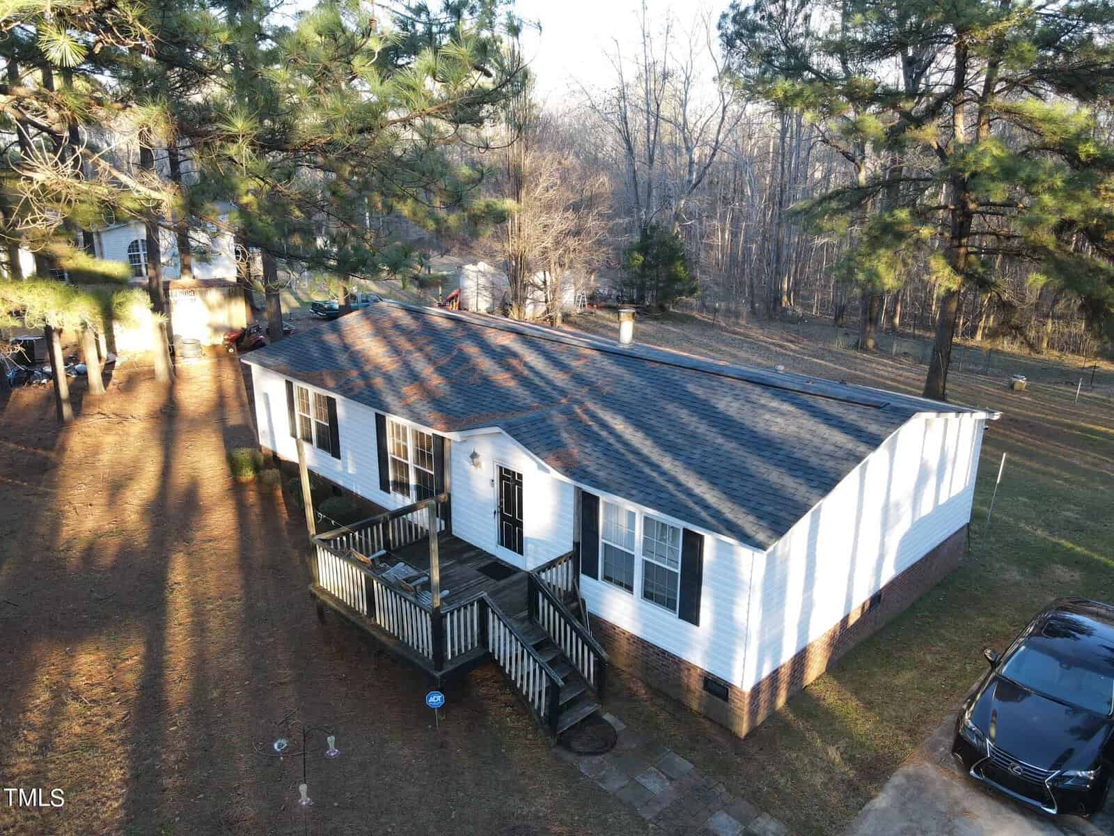 Aerial view of a white single-story house at 4038 Tyler Drive with a small front porch and dark roof, surrounded by large trees on a clear Oxford day. A car is parked nearby on the grassy lawn.