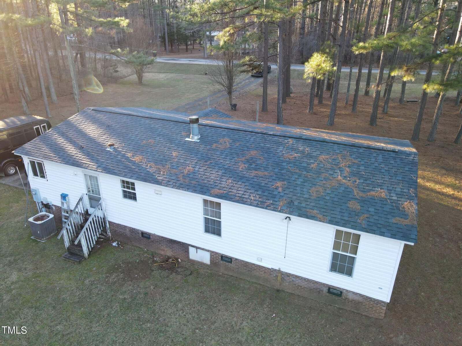 Aerial view of a single-story white house with a dark roof covered in pine needles. Surrounded by tall trees, the house has a small porch with a few steps leading to the yard. A parked van is visible next to the house.