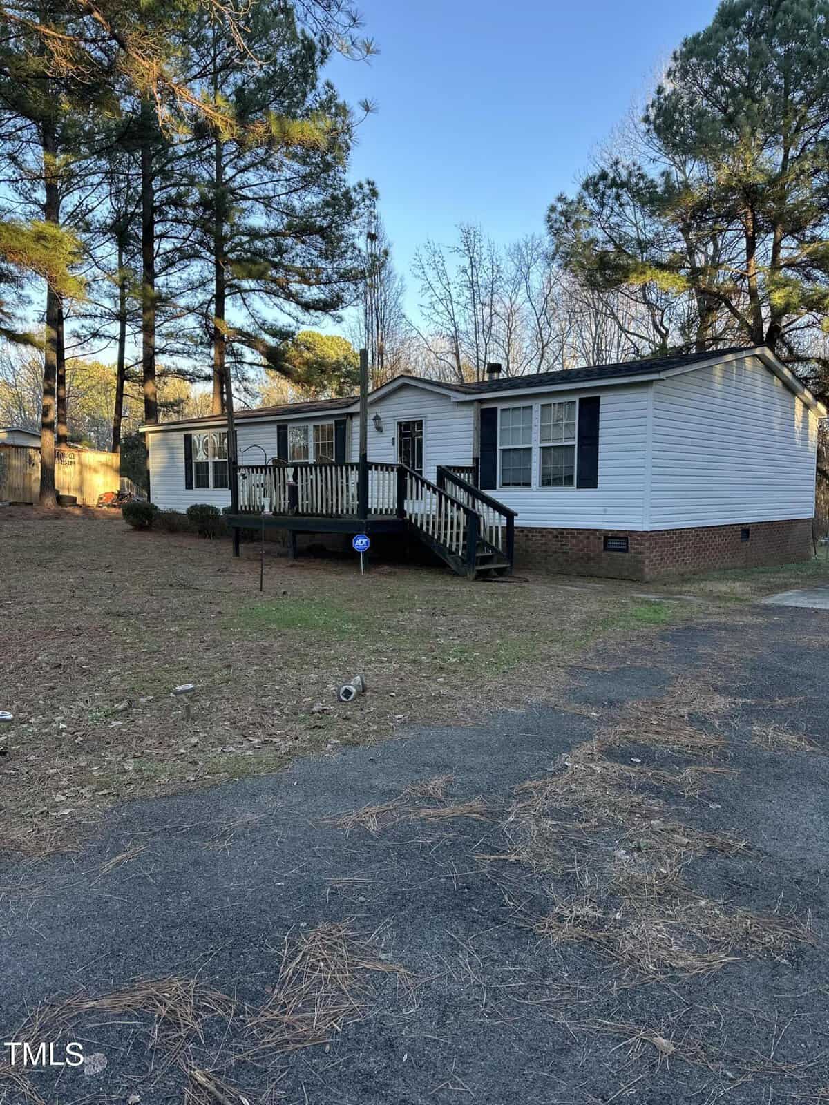 A single-story white house with a small front porch stands amidst tall trees. The yard is mostly grass with some fallen pine needles. A paved driveway leads up to the house. The sky is clear and blue.