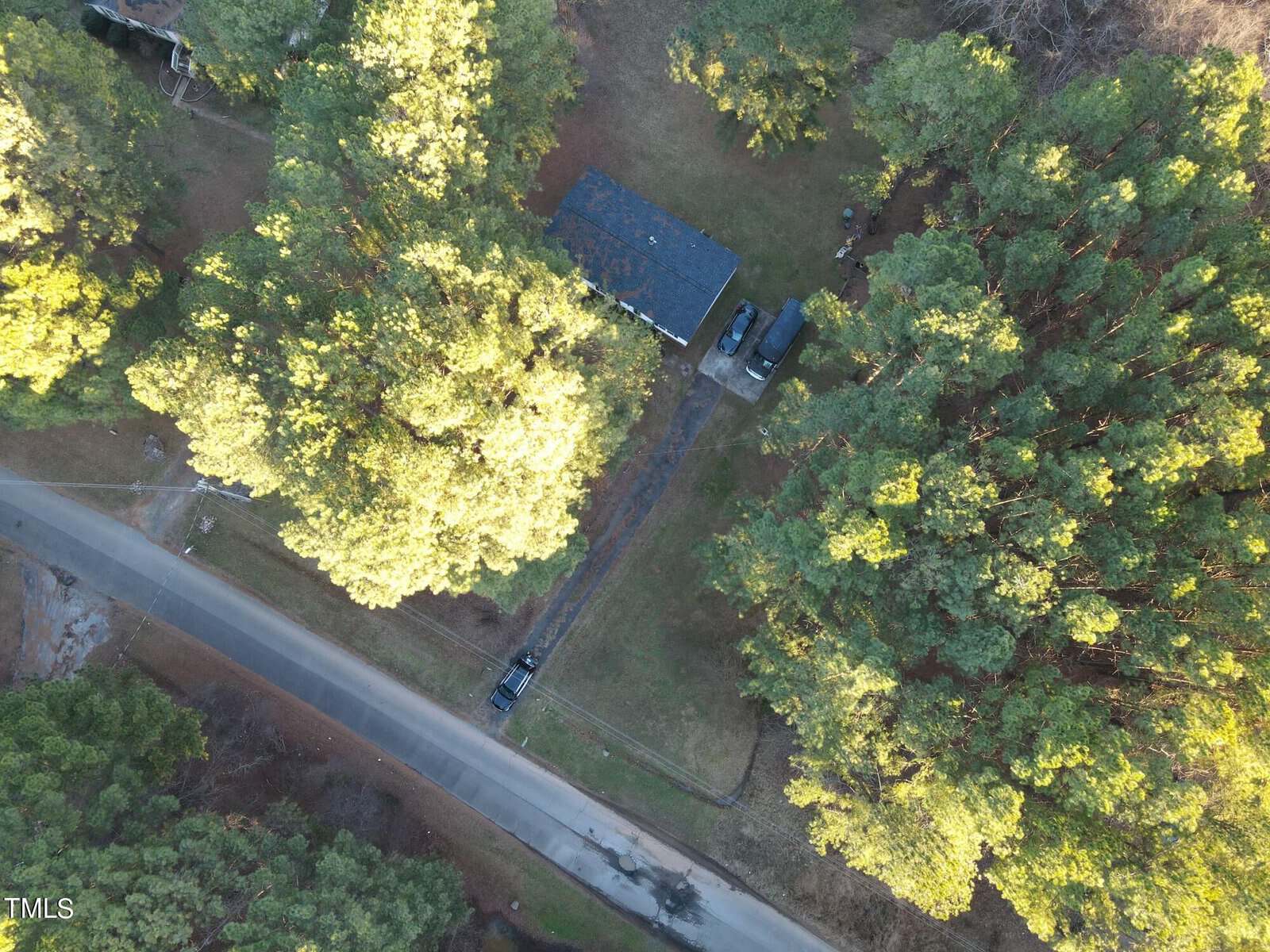 Aerial view of a house surrounded by dense green trees. A driveway leads from the house to a road, with two parked vehicles visible. The sunlight casts shadows through the trees, creating a serene natural setting.