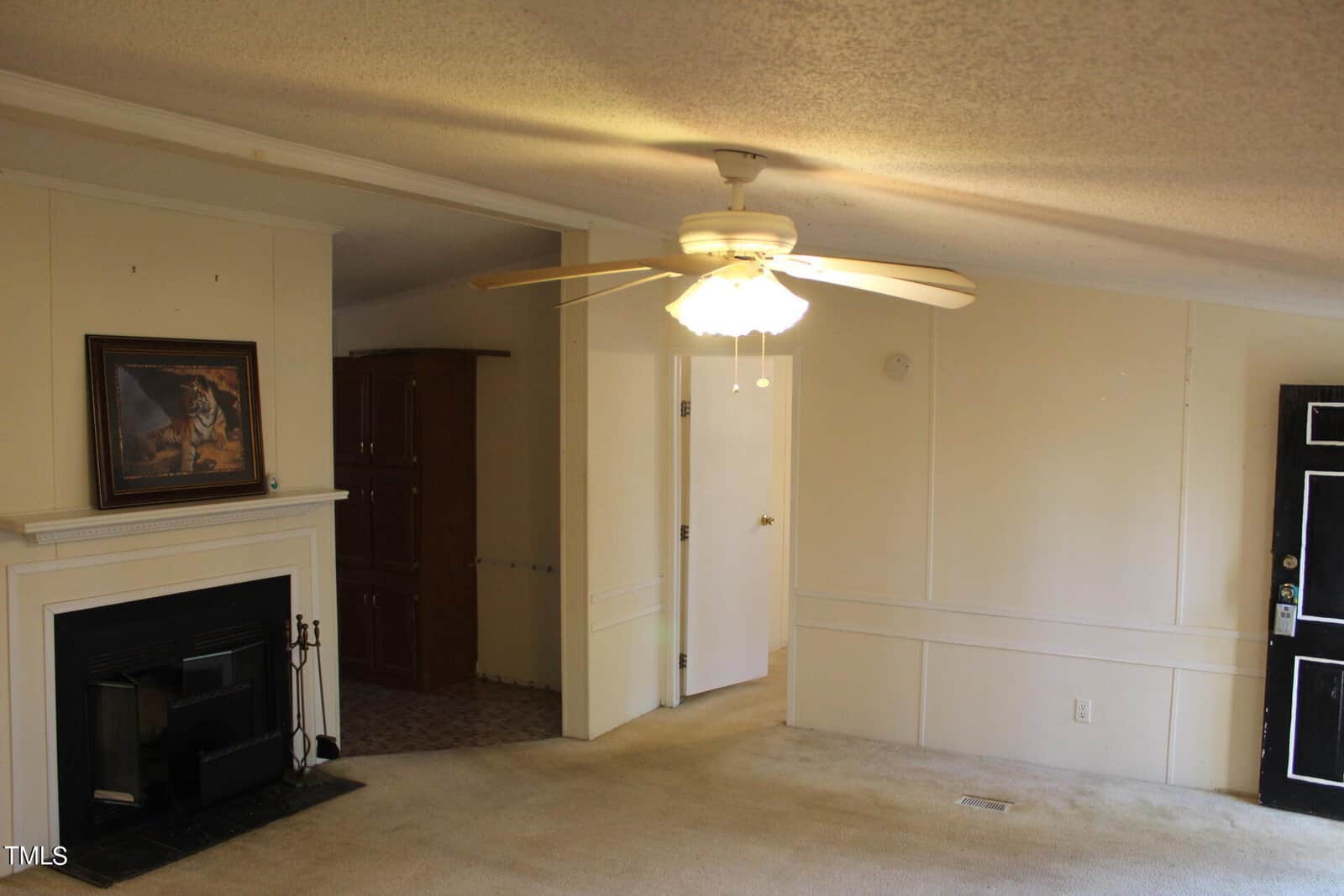 A living room with beige walls and carpet. There's a fireplace with a framed painting above it on the left. A ceiling fan with lights is centrally located. A door is partially open, revealing a glimpse of an adjacent room.