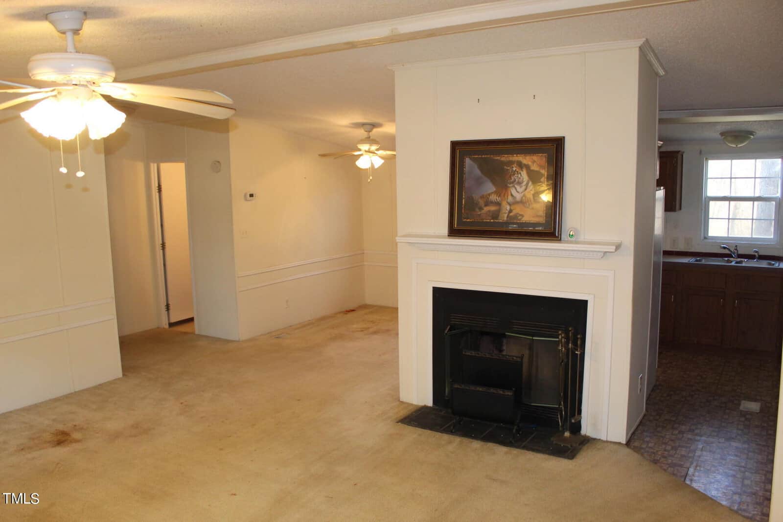 Interior of a home featuring a living area with beige carpet, a white ceiling with ceiling fans, and a fireplace adorned with a tiger painting. The room leads into a kitchen with wooden cabinetry and a window.