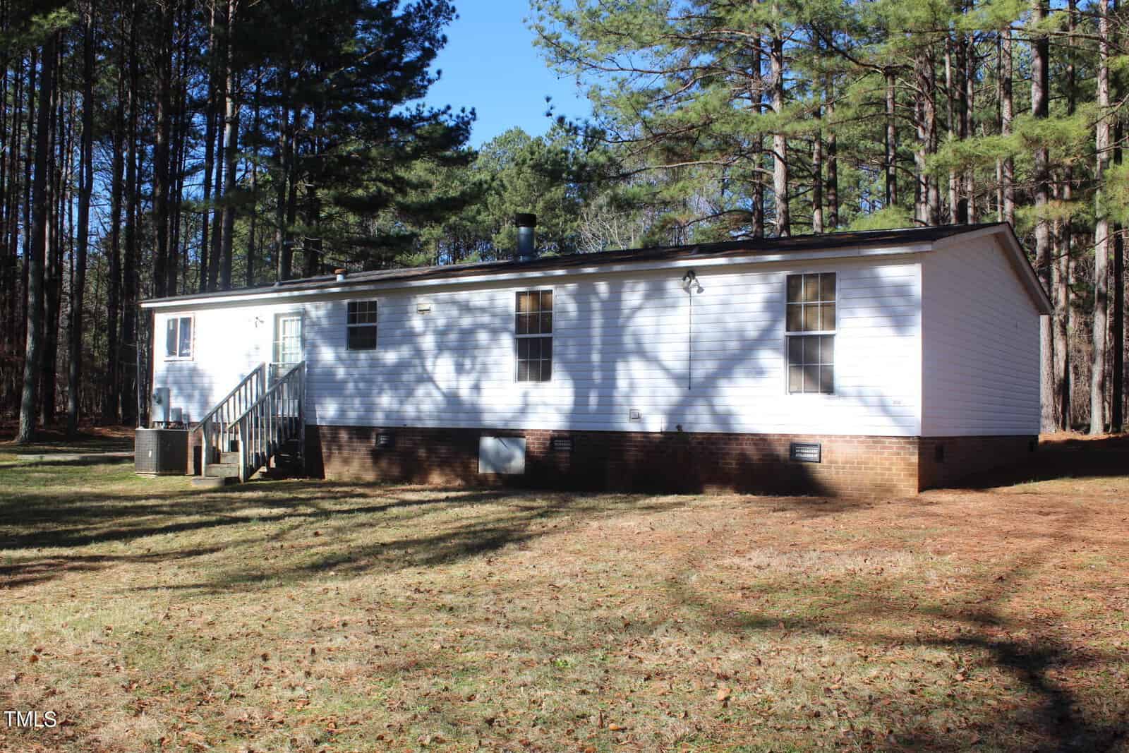A single-story white house with a brick foundation is surrounded by tall pine trees. Sunlight casts shadows on the grass and building. Steps lead up to a door on the side, and there are several windows visible on the house.