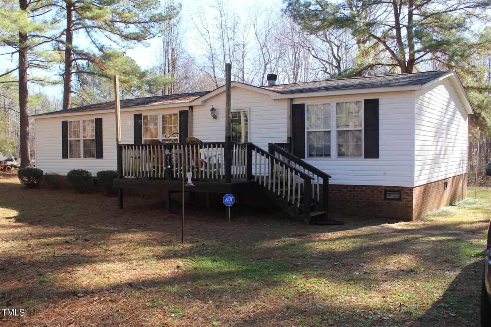 A white single-story house with a brick foundation and a small front porch is situated in a wooded area. The porch has a railing and steps leading down. Tall pine trees surround the house, and a security sign is visible in the yard.