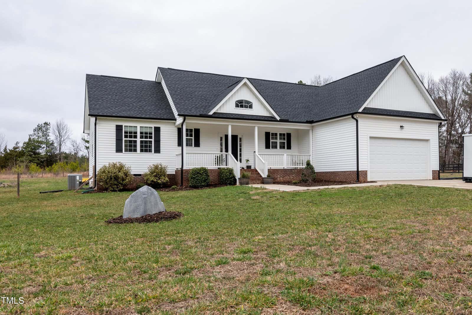 A white, single-story house at 4081 Old Nc 75, with a dark roof and porch, features a two-car garage set on a grassy lawn. Steps ascend to the porch, and a large rock stands prominently in the foreground. Trees are visible against an overcast sky.