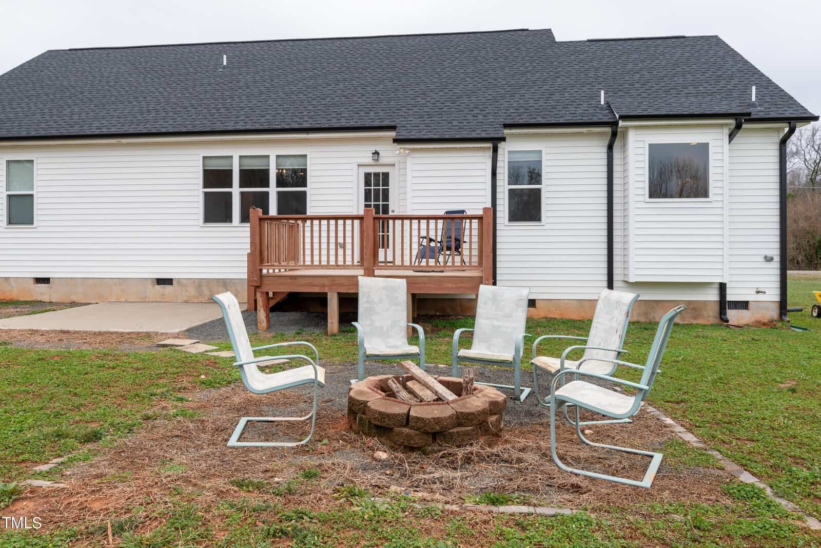 A white house with a small wooden deck and a sliding glass door. In the backyard, there are five patio chairs arranged around a circular stone fire pit on a patch of grass. The sky is overcast.