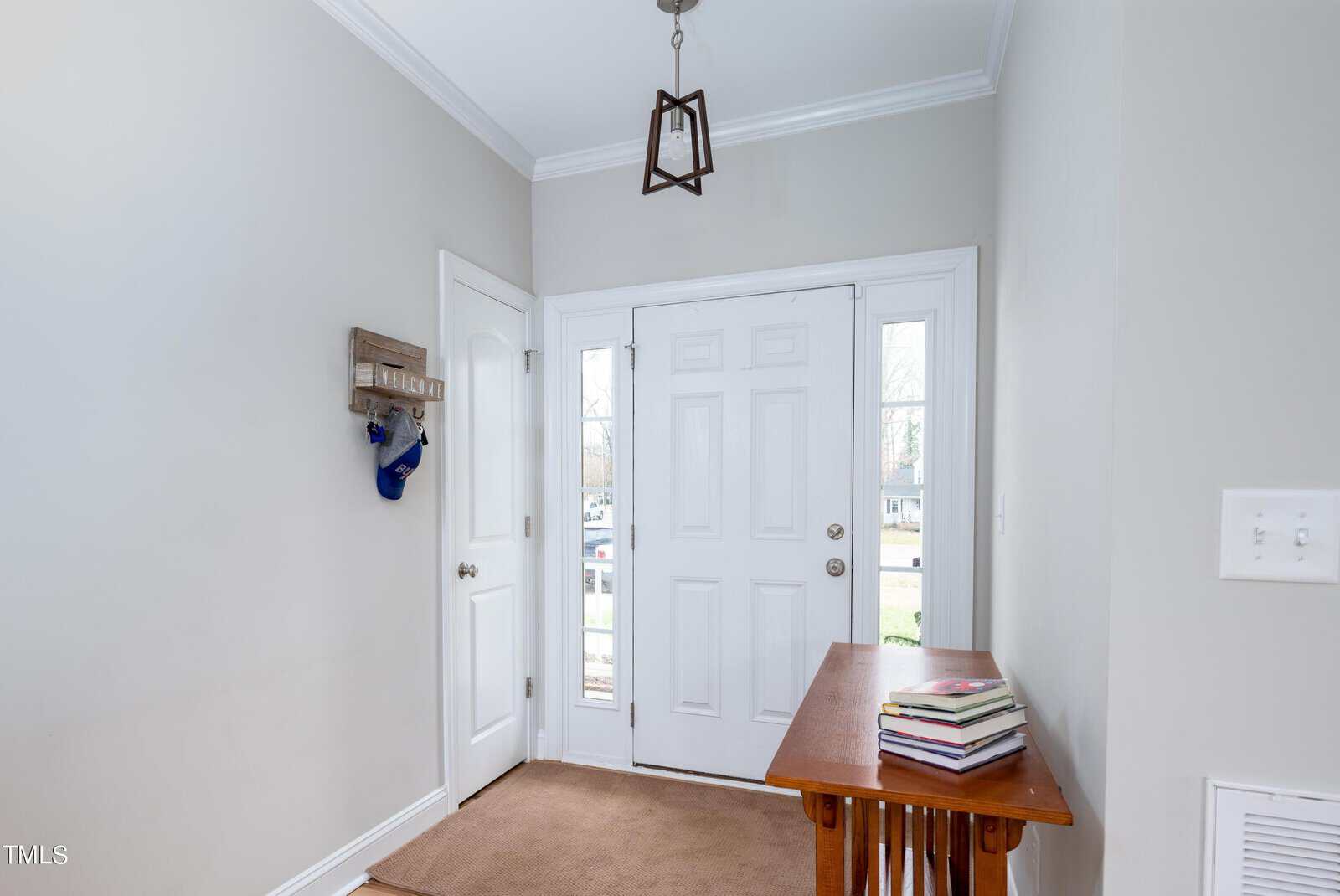 A bright entryway with white double doors. A small wooden table on the right holds stacked books. A wall-mounted hook with keys and a baseball cap is on the left. The floor is carpeted, and a modern pendant light hangs overhead.