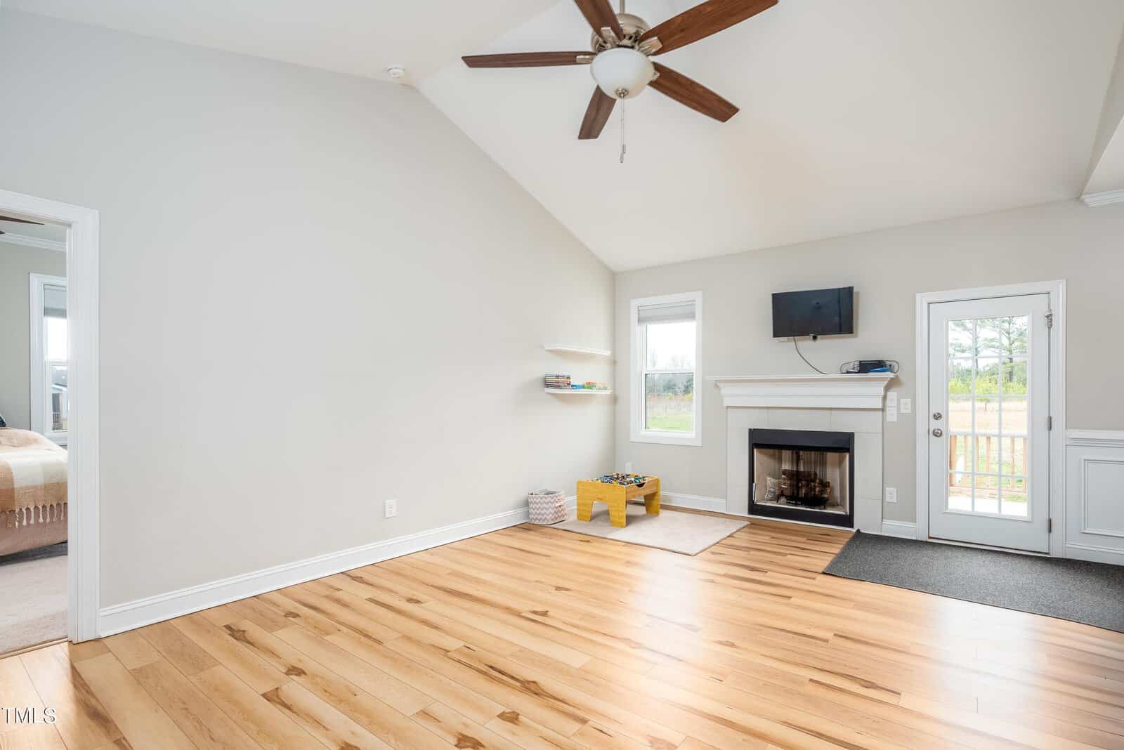 A spacious room with light wooden flooring, a ceiling fan, and a fireplace. There's a wall-mounted TV above the fireplace, a small play area with toys, and a door leading to an outside view. A doorway on the left leads to another room.
