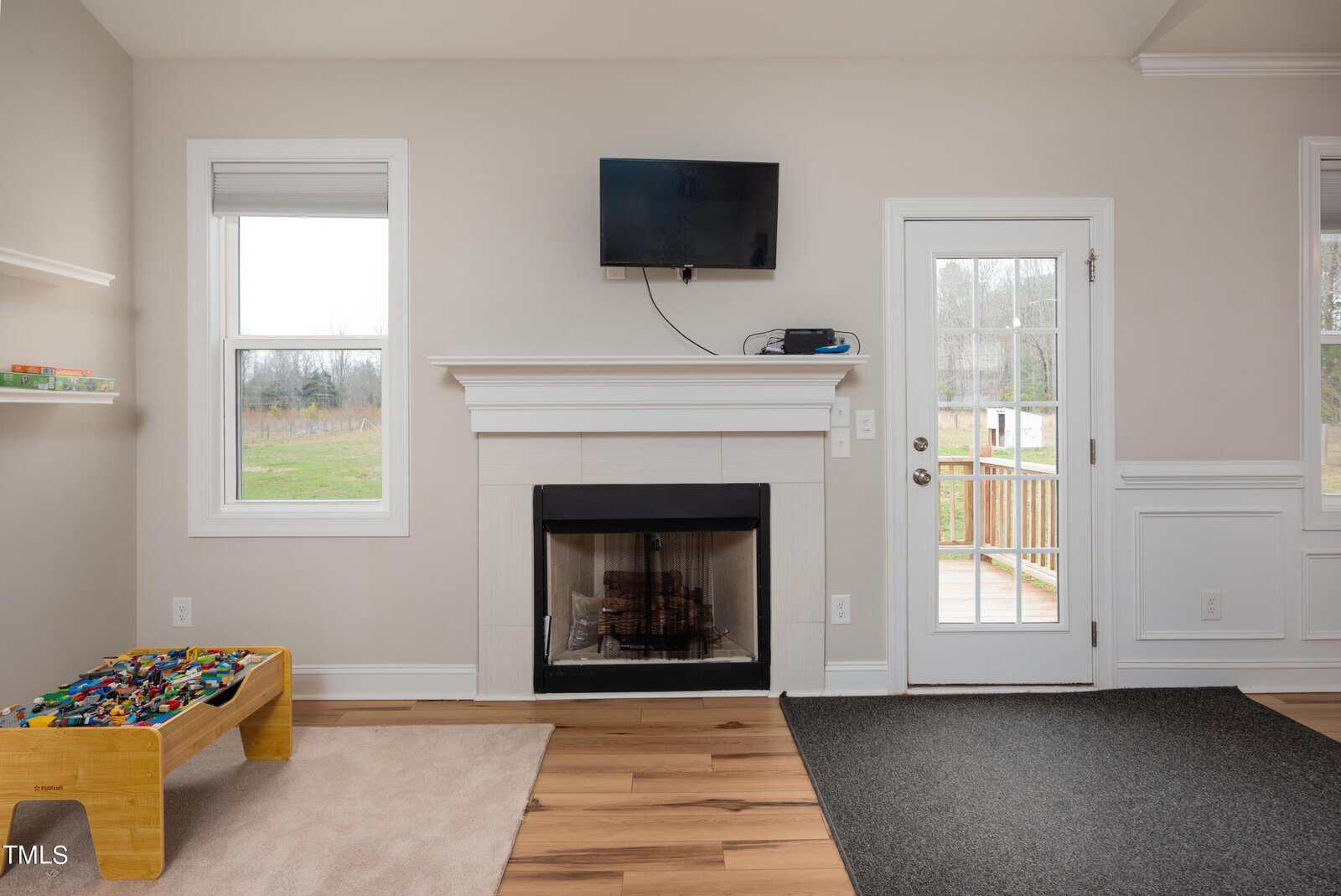 Living room with a mounted TV above a white fireplace, a glass door leading to a deck, large windows, and a play table with toys. The space features light-colored walls and wood flooring.