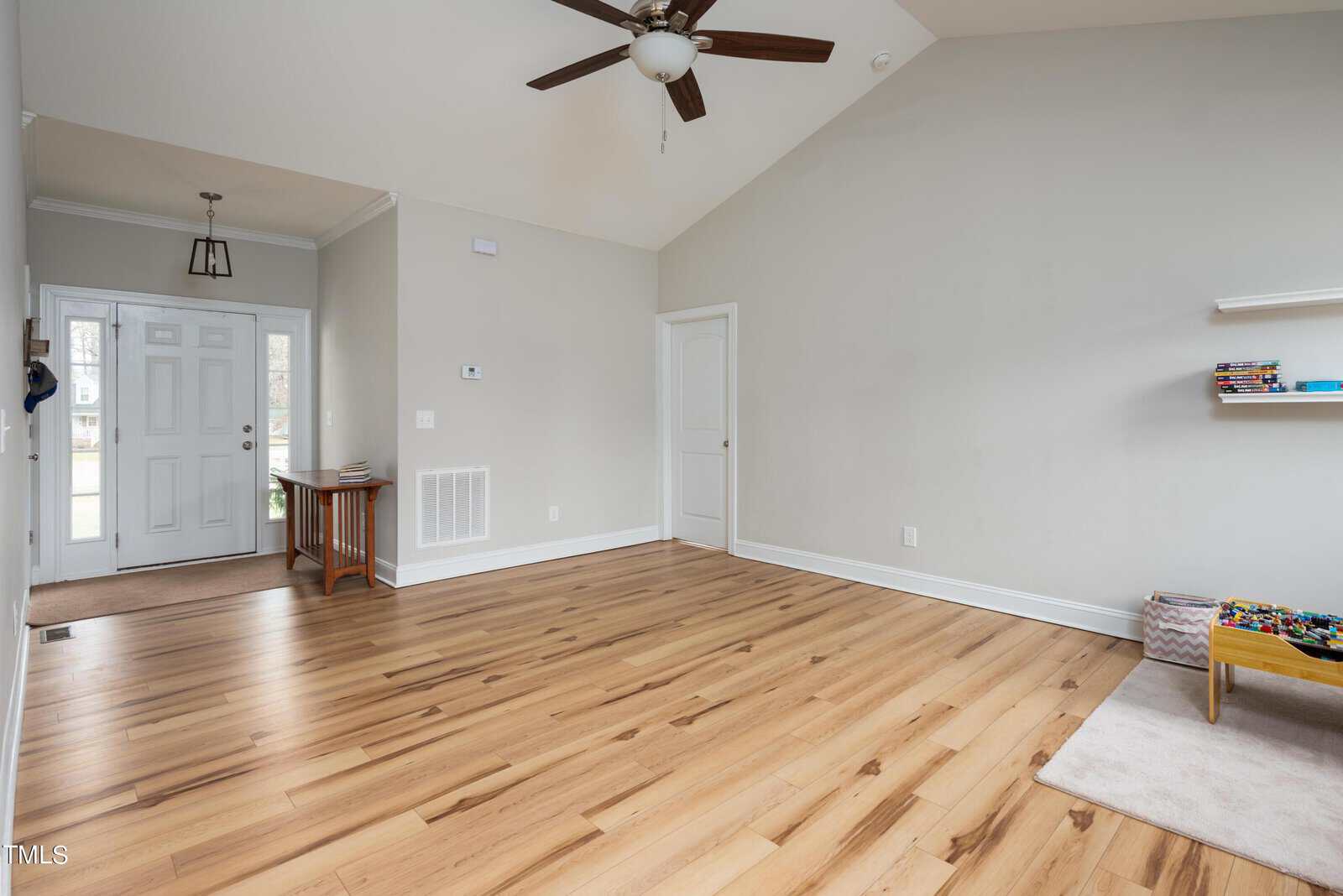 A spacious, well-lit living room with light wood flooring and a vaulted ceiling. The room features a ceiling fan, a small rug with toys, a shelf with books, and a view towards a doorway with a small wooden table. Walls are painted in a light color.