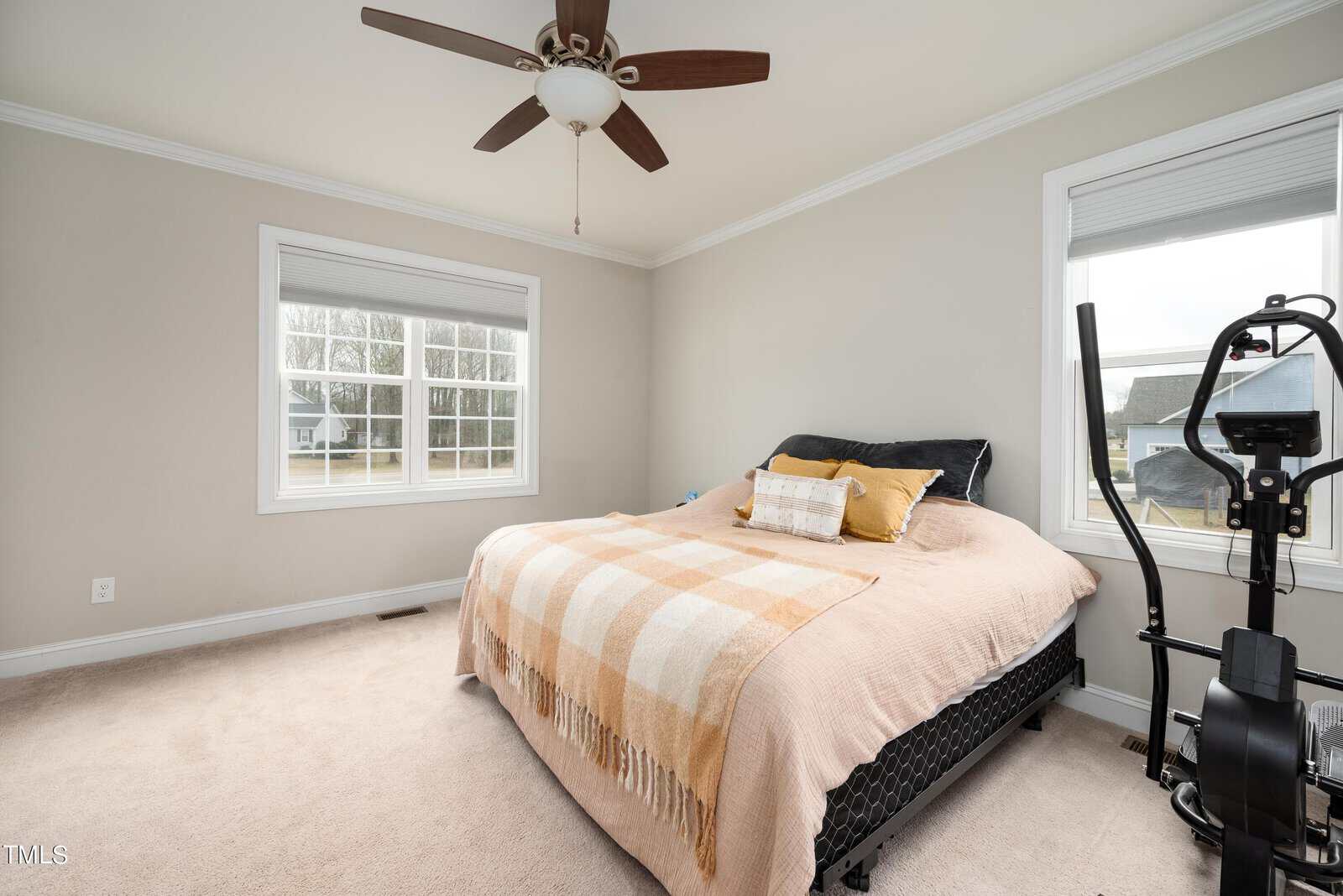 A bedroom with a double bed covered in beige and orange blankets and cushions. There's a ceiling fan, an exercise machine, and two large windows letting in natural light. The room has light-colored walls and carpet.
