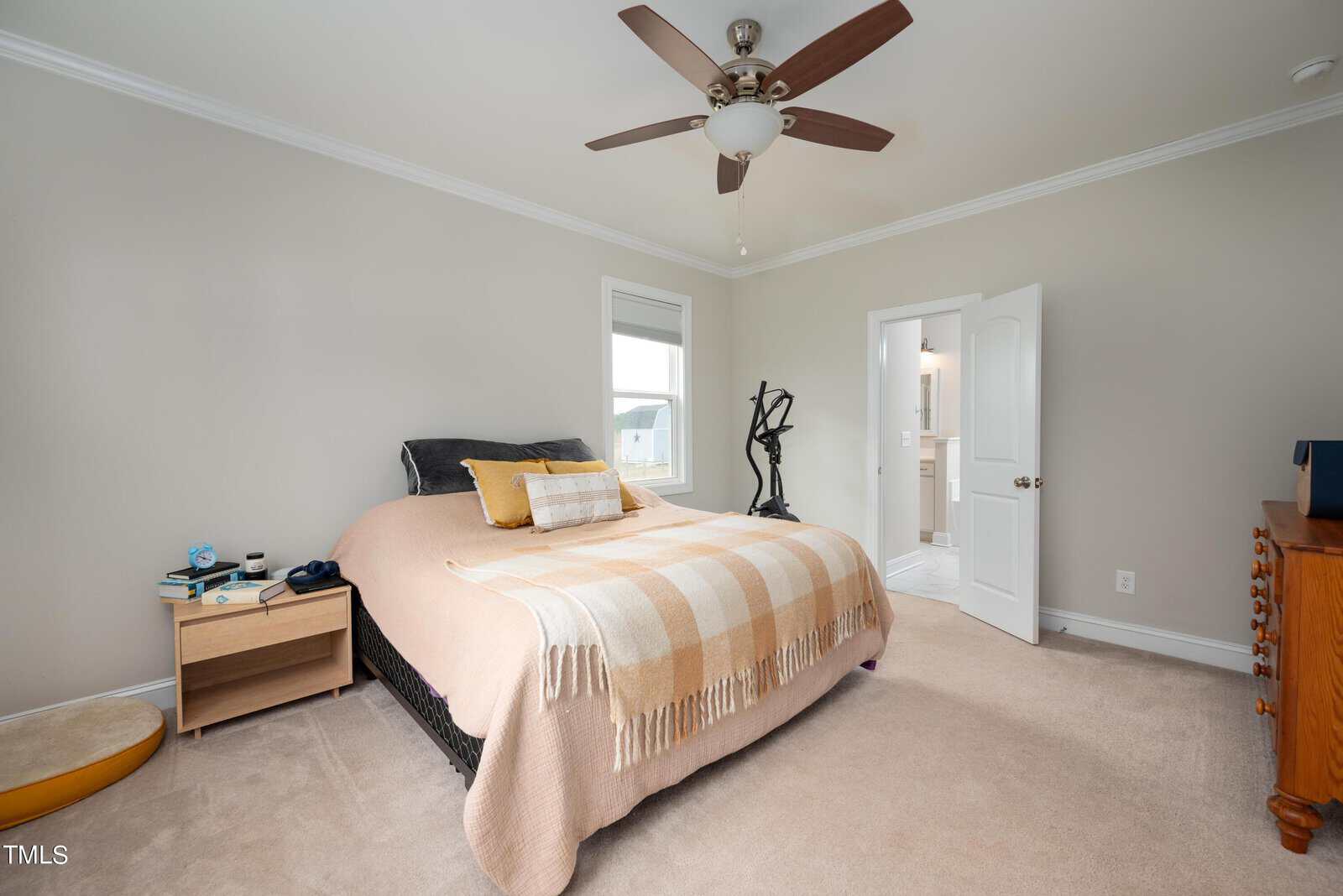 A bedroom with a large bed covered in a beige plaid blanket and yellow pillow. A ceiling fan is above. The room has a nightstand with items, a window, and an open door leading to a bathroom. A dresser and decorative object are visible.