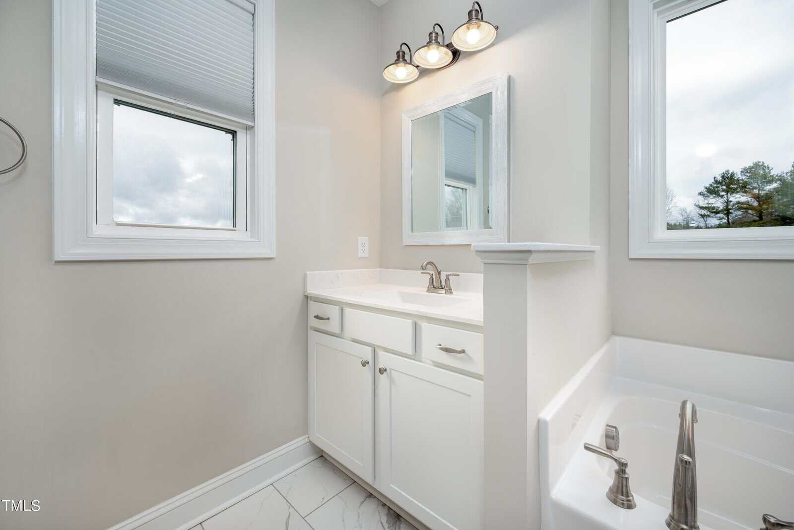 A bright bathroom with a white vanity, marble countertop, and silver faucet. Above the vanity is a rectangular mirror with three overhead lights. The room has two windows, and part of a white bathtub is visible on the right.
