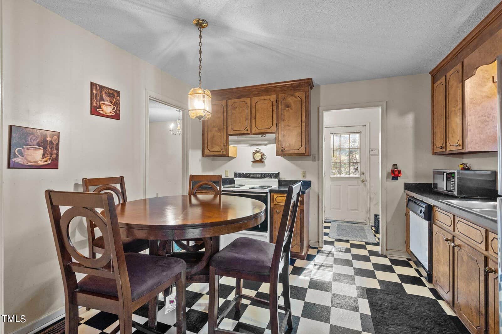 A kitchen with a black and white checkered floor, wooden dining table set with four chairs, wooden cabinets, and appliances including a microwave and stove. a pendant light hangs above the table.