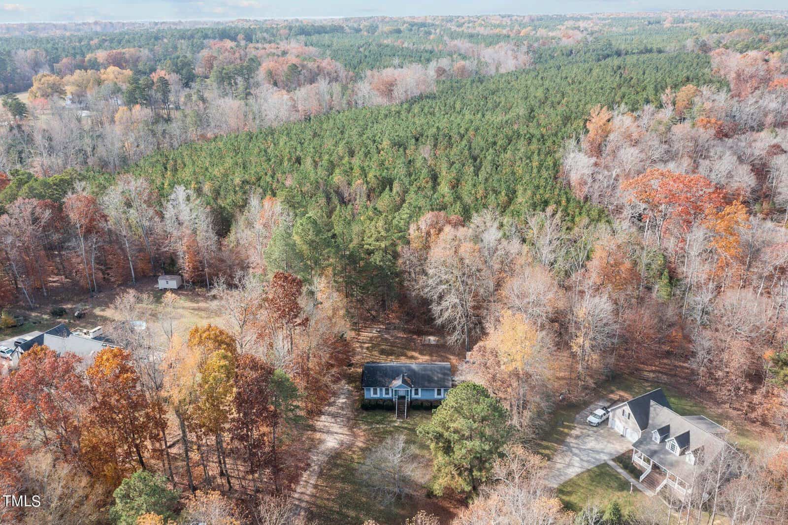 Aerial view of two houses surrounded by woods displaying autumn colors, one house is single-story and the other is larger with multiple roofs. the landscape is a mix of evergreen and deciduous trees.